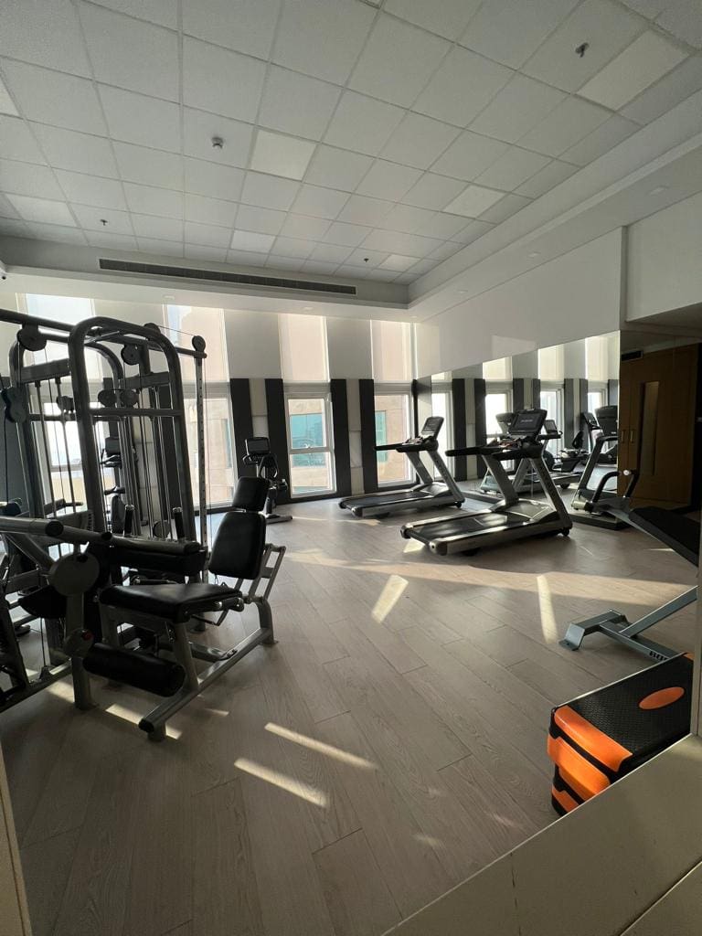 A well-lit gym in the Juffair apartment complex features various exercise equipment, including treadmills, a multi-station gym machine, and a step platform. Large windows allow natural light to brighten the space, making it an ideal spot for residents to work out. Available for rent now!