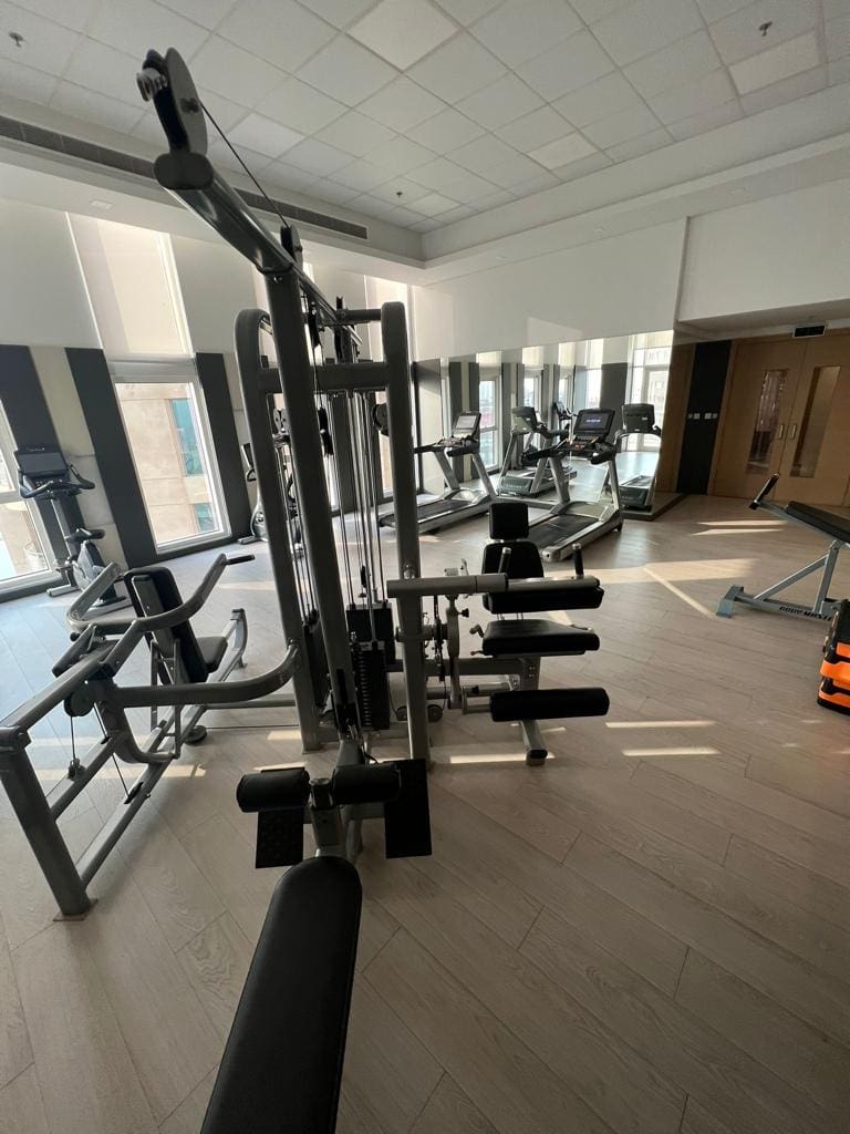 A gym in Juffair with various exercise equipment, including a cable machine, treadmills, benches, and weights, illuminated by natural and artificial light. Perfect for residents looking to rent an apartment with top-notch fitness facilities.