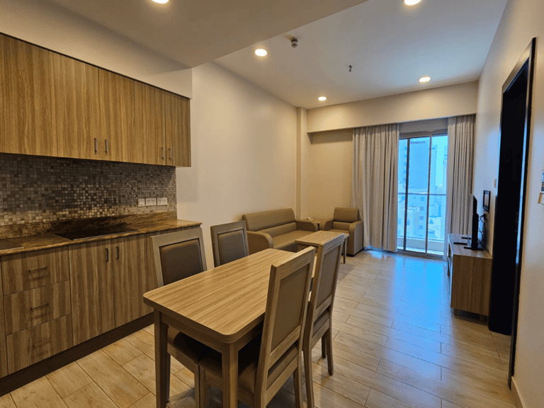 A modern, furnished flat for rent in Juffair features a living area with a kitchenette, dining table, chairs, sofa, armchair, wall-mounted TV, and a glass door leading to a balcony. The room boasts wooden flooring and ample lighting.