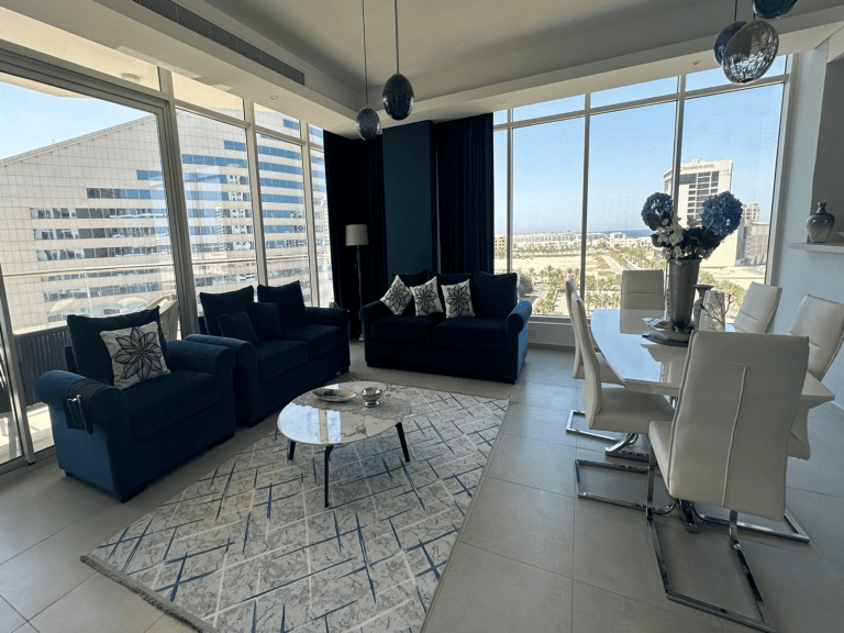 A modern living and dining area with blue sofas, a white and blue rug, a white dining table with white chairs, large windows offering an urban view, and a glass door opening to a balcony. This stylish flat for rent in Amwaj is perfect for contemporary living.