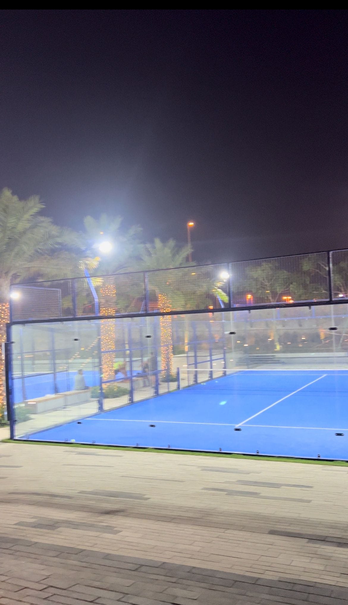 A brightly lit outdoor blue padel court at night, surrounded by palm trees wrapped in string lights, feels like an auto draft escape into a tropical oasis.