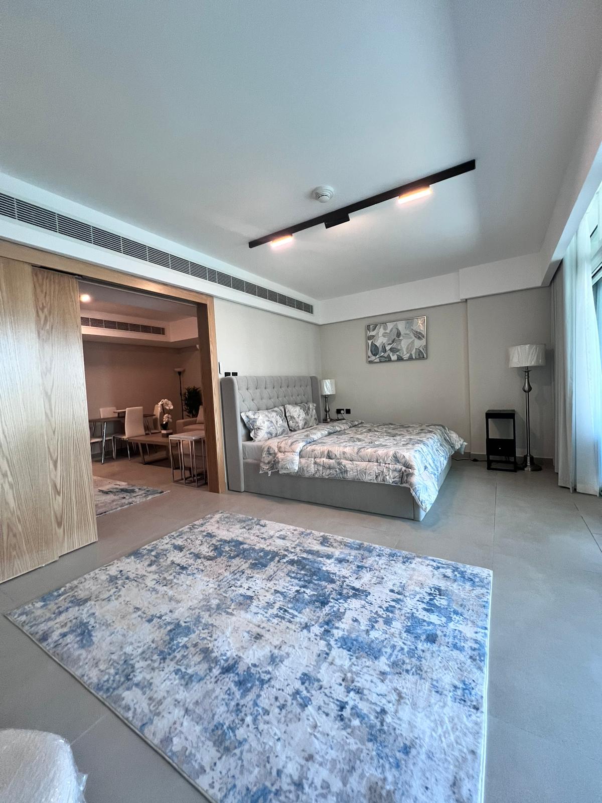 Modern bedroom with a large patterned rug, a light gray upholstered bed, and minimalistic furniture. An open doorway reveals a dining area in the background. Bright natural light fills the room, creating an atmosphere that feels effortlessly styled like an Auto Draft masterpiece.