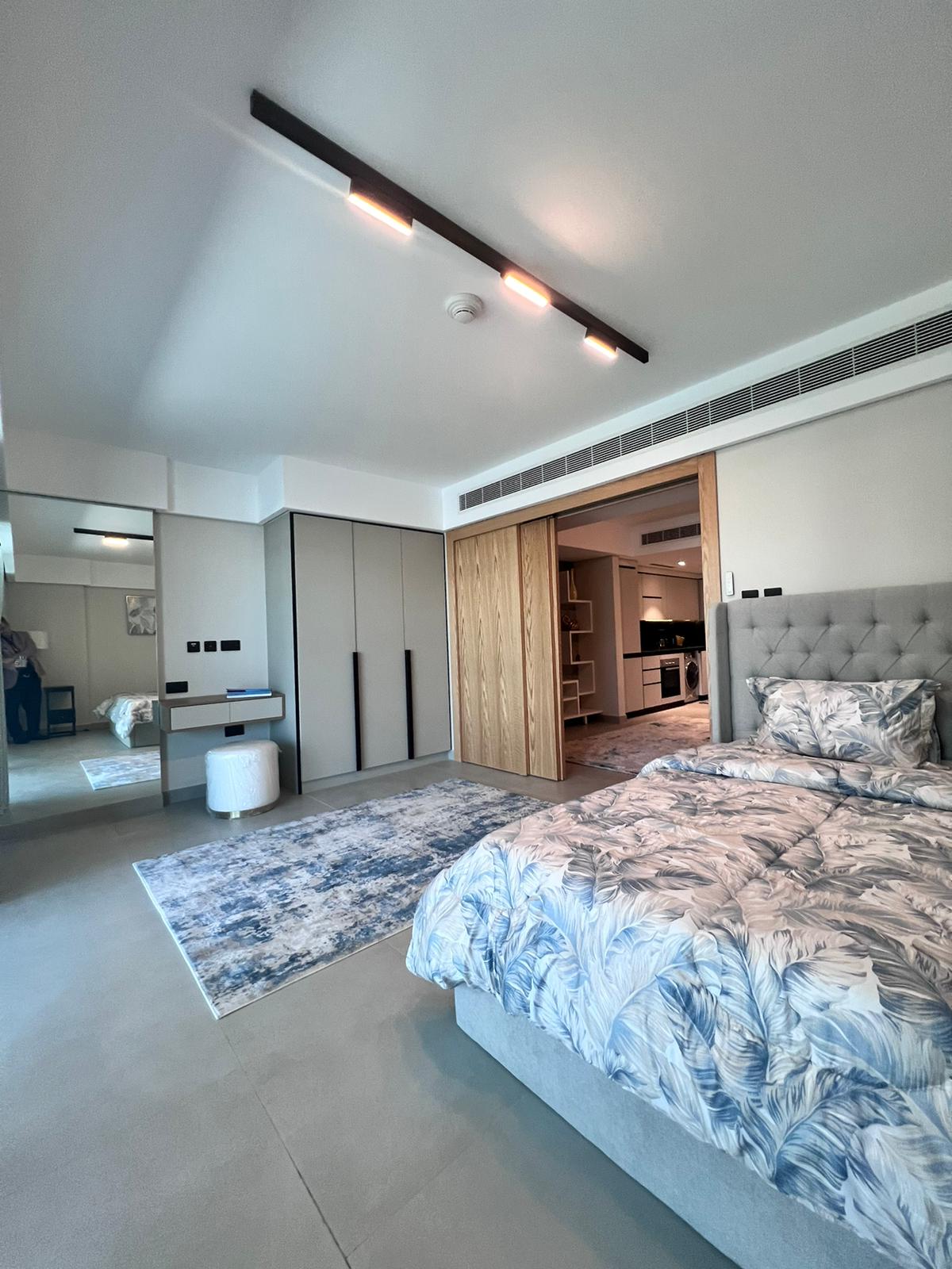 A modern bedroom with a grey upholstered bed, floral bedding, a large mirror, and sliding wooden closet doors. A blue rug is on the floor, and the room has recessed lighting.