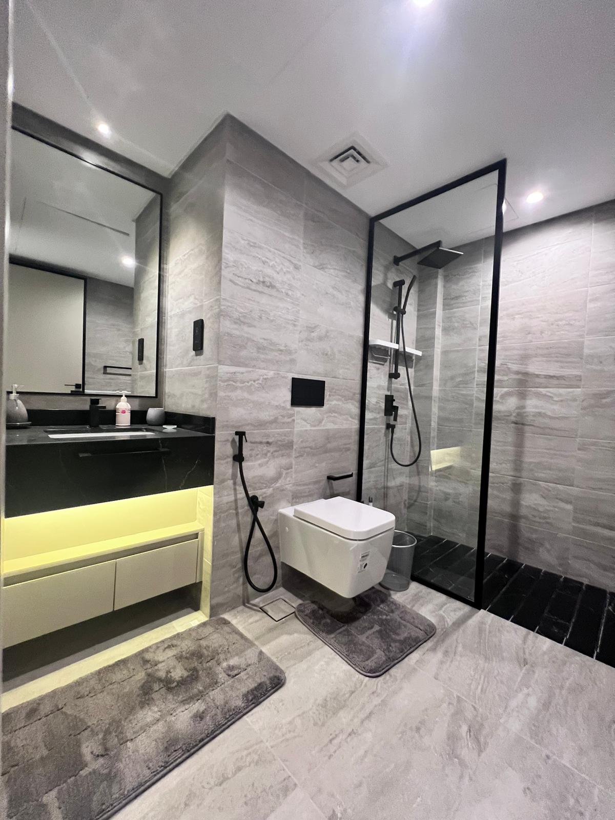 Modern bathroom with grey tiles, a wall-mounted toilet, black fixtures, and a walk-in glass shower. An illuminated vanity with a large mirror adds elegance, complemented by two grey floor mats. This auto-draft design seamlessly blends function and style for a contemporary feel.