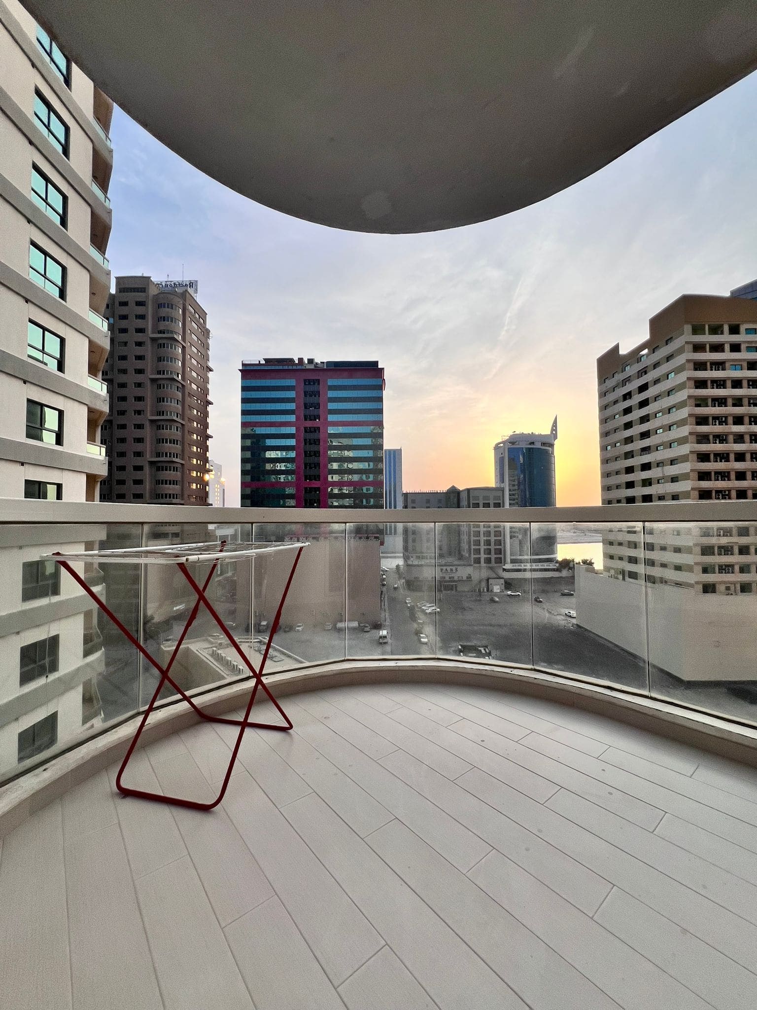 A curved balcony with a red metal stand overlooks the Juffair cityscape, featuring several tall buildings under a partly cloudy sky at sunset—an ideal apartment for rent.