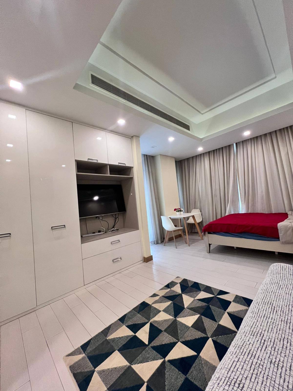 A modern bedroom in a Juffair apartment features white-themed decor, a large wardrobe, a bed with a red blanket, a small desk with chairs, and a TV. The space is enhanced by ample lighting and a geometric-patterned rug, making it an inviting option for rent.