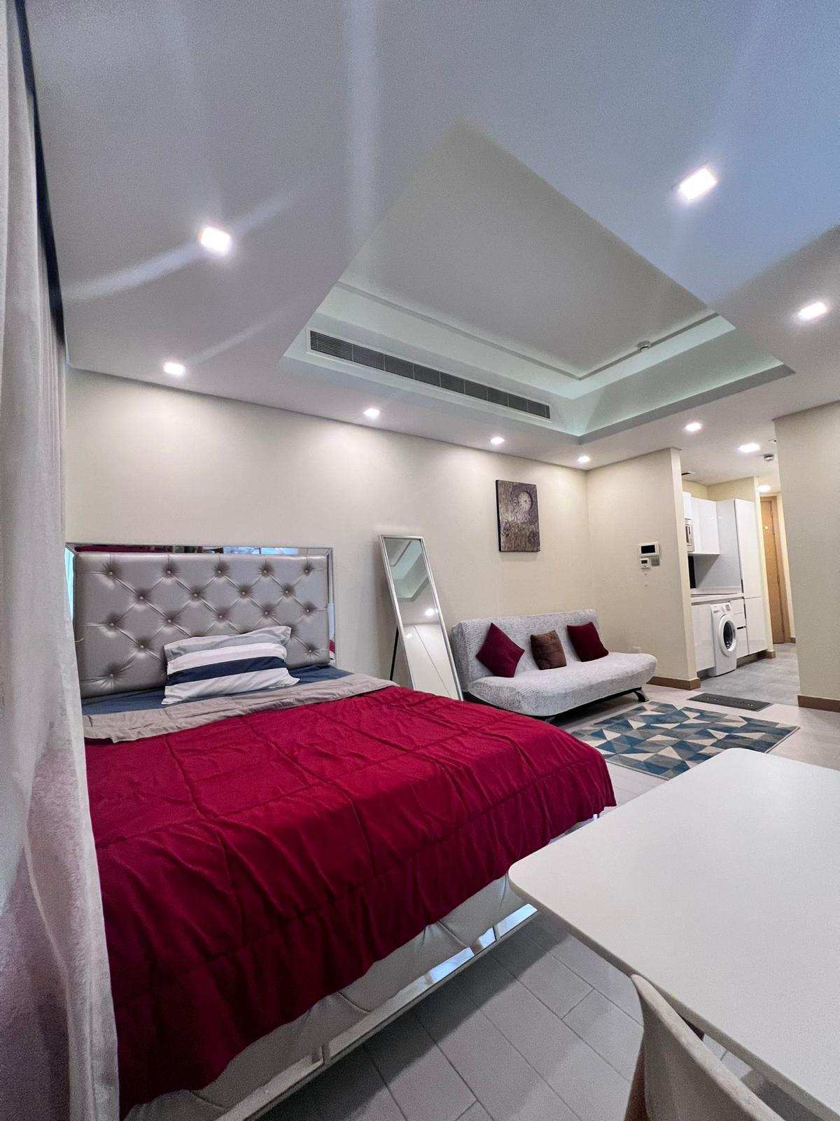 A modern Juffair studio apartment for rent featuring a bed with a red comforter, a white sofa with red pillows, a wall-mounted mirror, a small kitchen area with appliances, and ceiling recessed lighting.