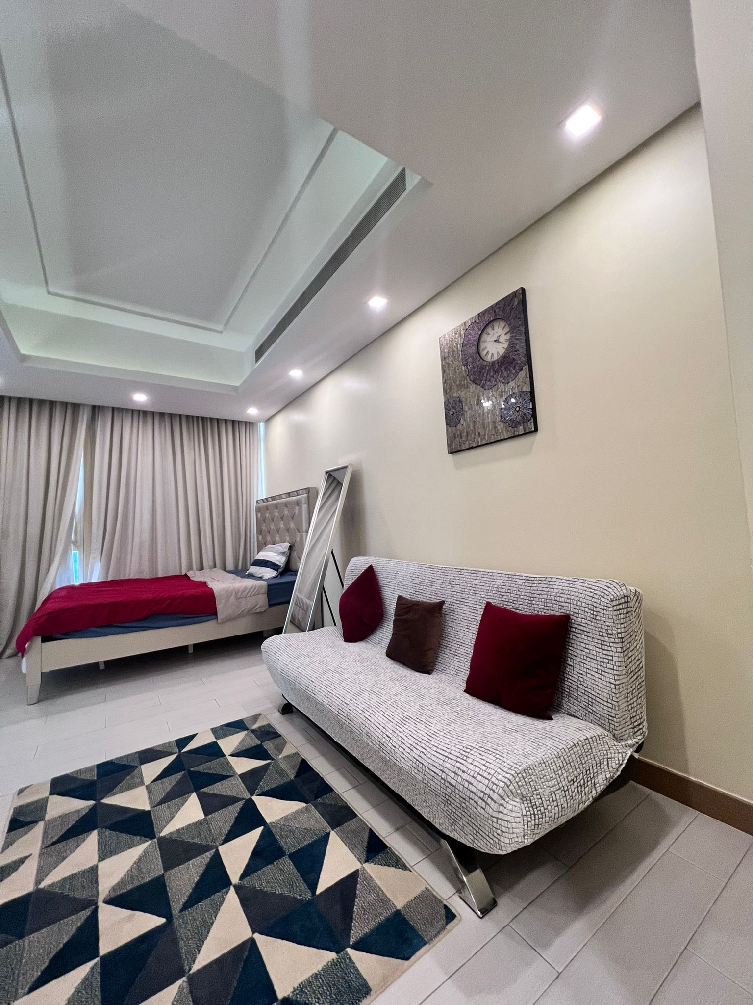 A bedroom in an apartment for rent in the Juffair area features a bed, patterned rug, and a cozy couch with cushions. The wall showcases a clock-themed art piece, while a large mirror behind the couch adds depth to the space. The room is well-lit with ceiling lights.