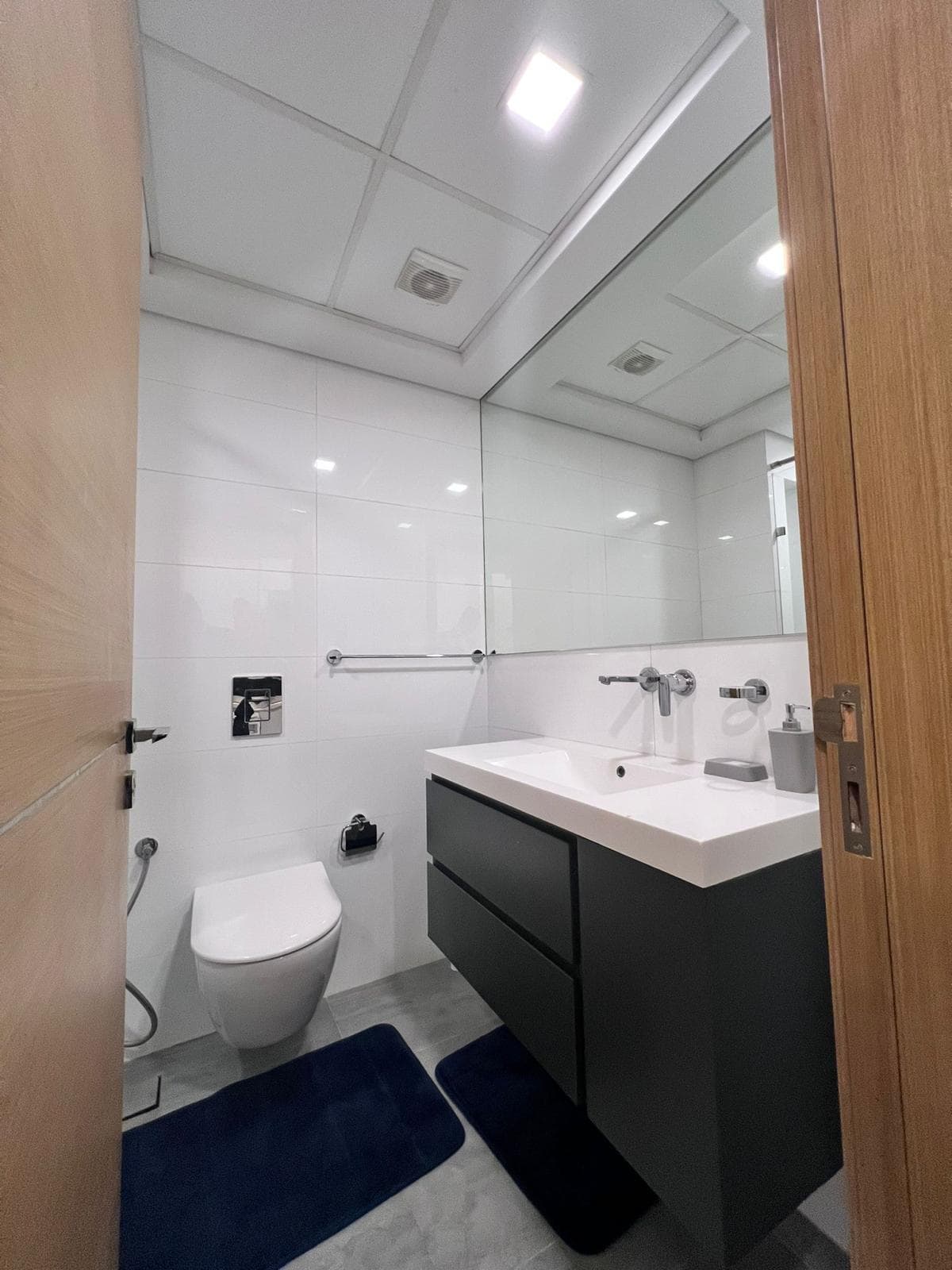 A compact bathroom in a Juffair apartment features a wall-mounted toilet, a large mirror above a sink with storage drawers underneath, and two dark blue rugs on the floor. Ideal for renters seeking convenience and style.