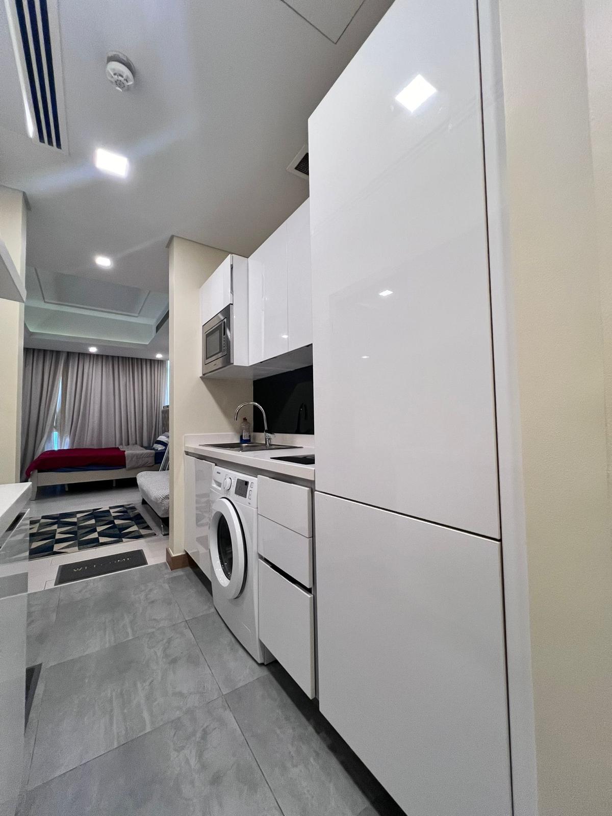 A modern studio apartment in Juffair is available for rent, featuring a compact kitchen with white cabinets, a microwave, and a washing machine. The small living area includes both a bed and couch for your convenience.