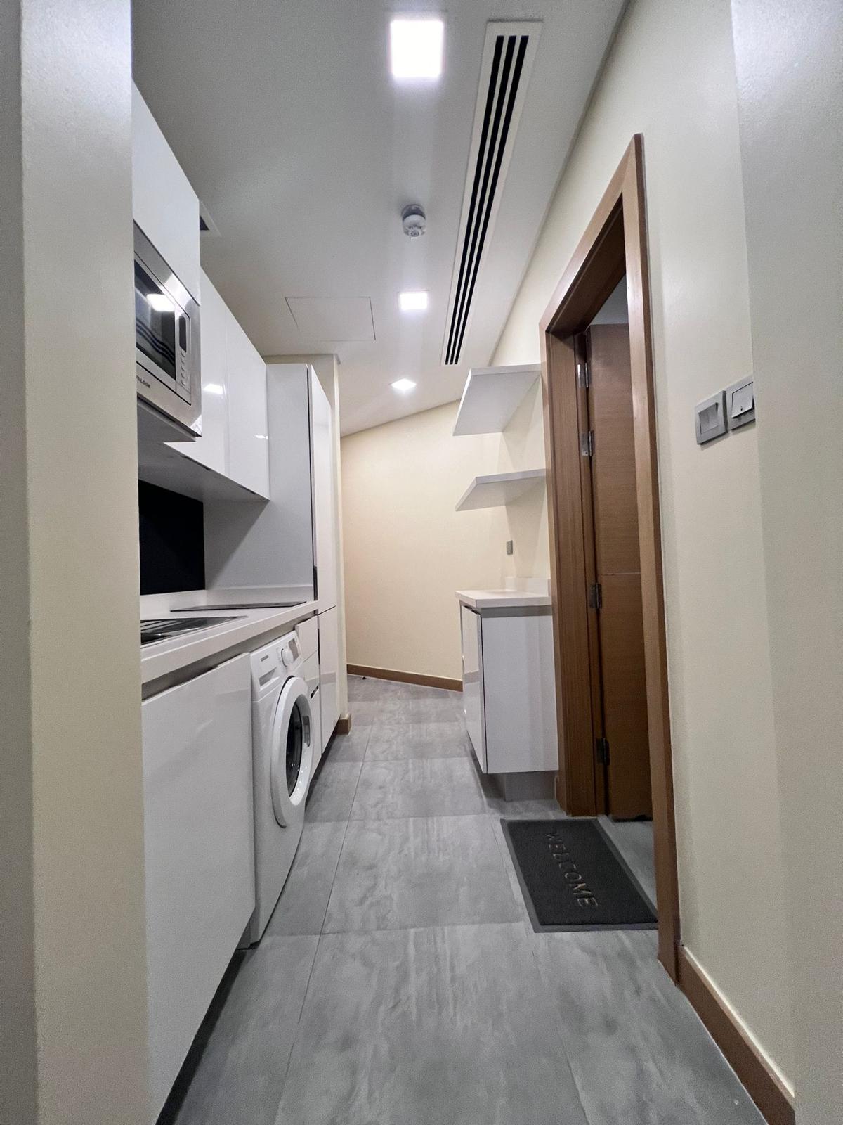 A narrow kitchen with white cabinets, a microwave, a washing machine, and shelves in an apartment for rent in Juffair. The floor is gray tile, the walls are light-colored, and a wooden door is visible on the right.