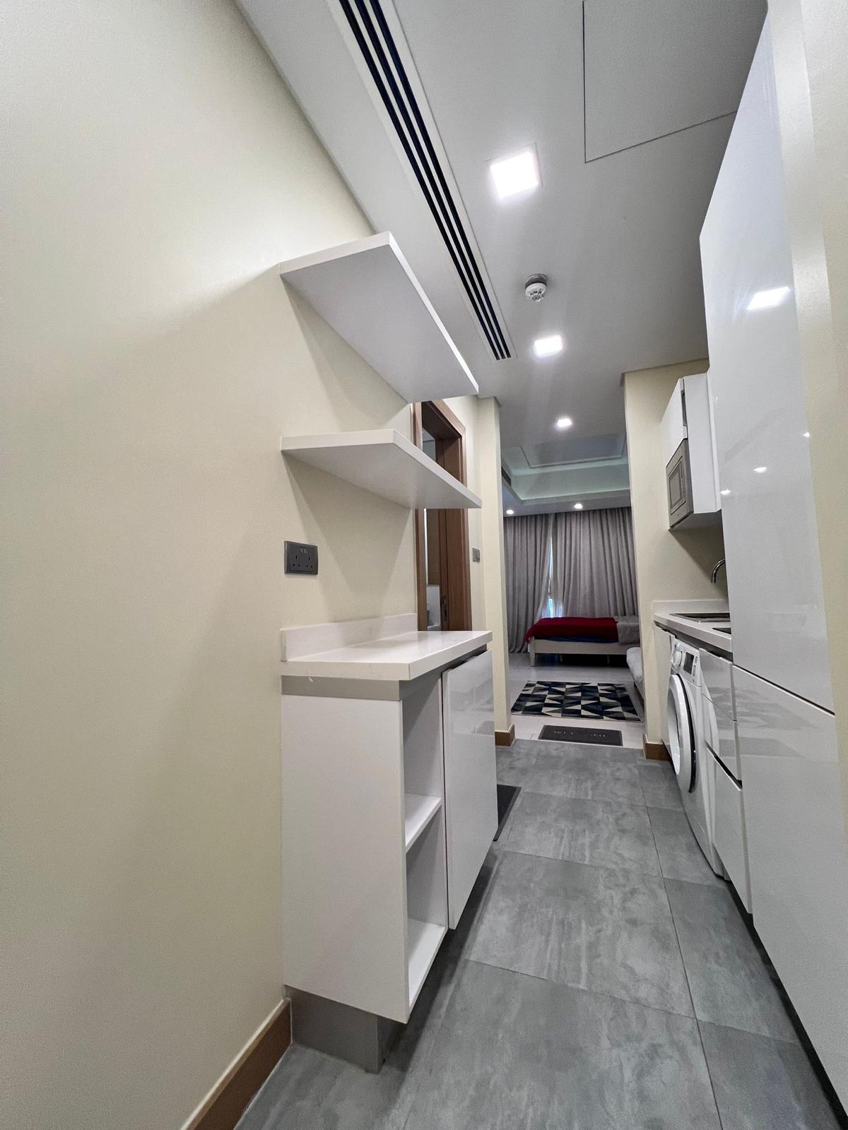 A modern, compact kitchen with white cabinetry, built-in appliances including a washing machine and microwave, shelves, and a view into the living area featuring a bed and a window with curtains. This stylish apartment in Juffair is now available for rent.