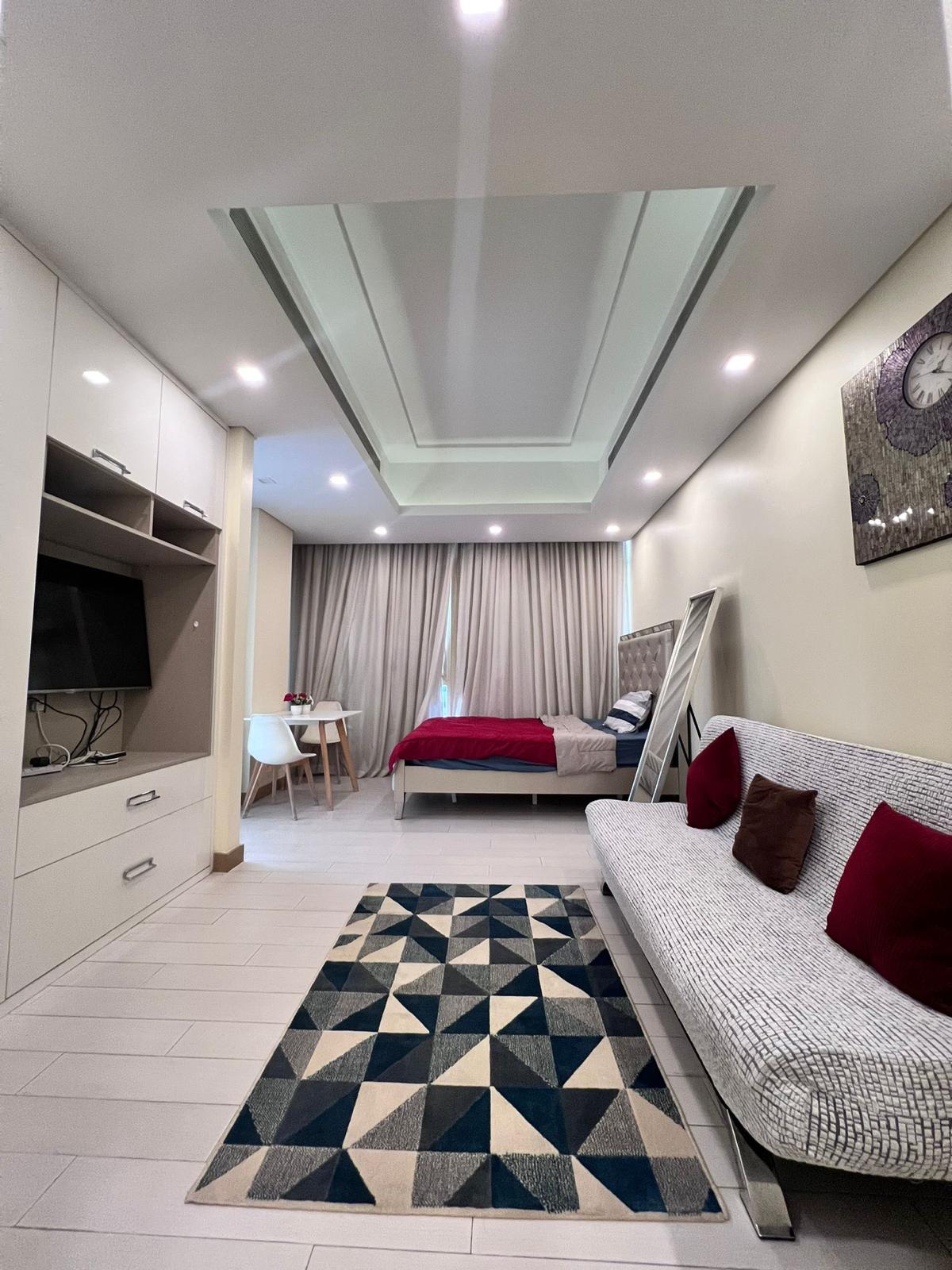 A modern studio apartment for rent in the Juffair area with a bed, a sofa, a TV, a desk, and a geometric-patterned rug. The room features recessed ceiling lights, large curtains, and minimalist decor.