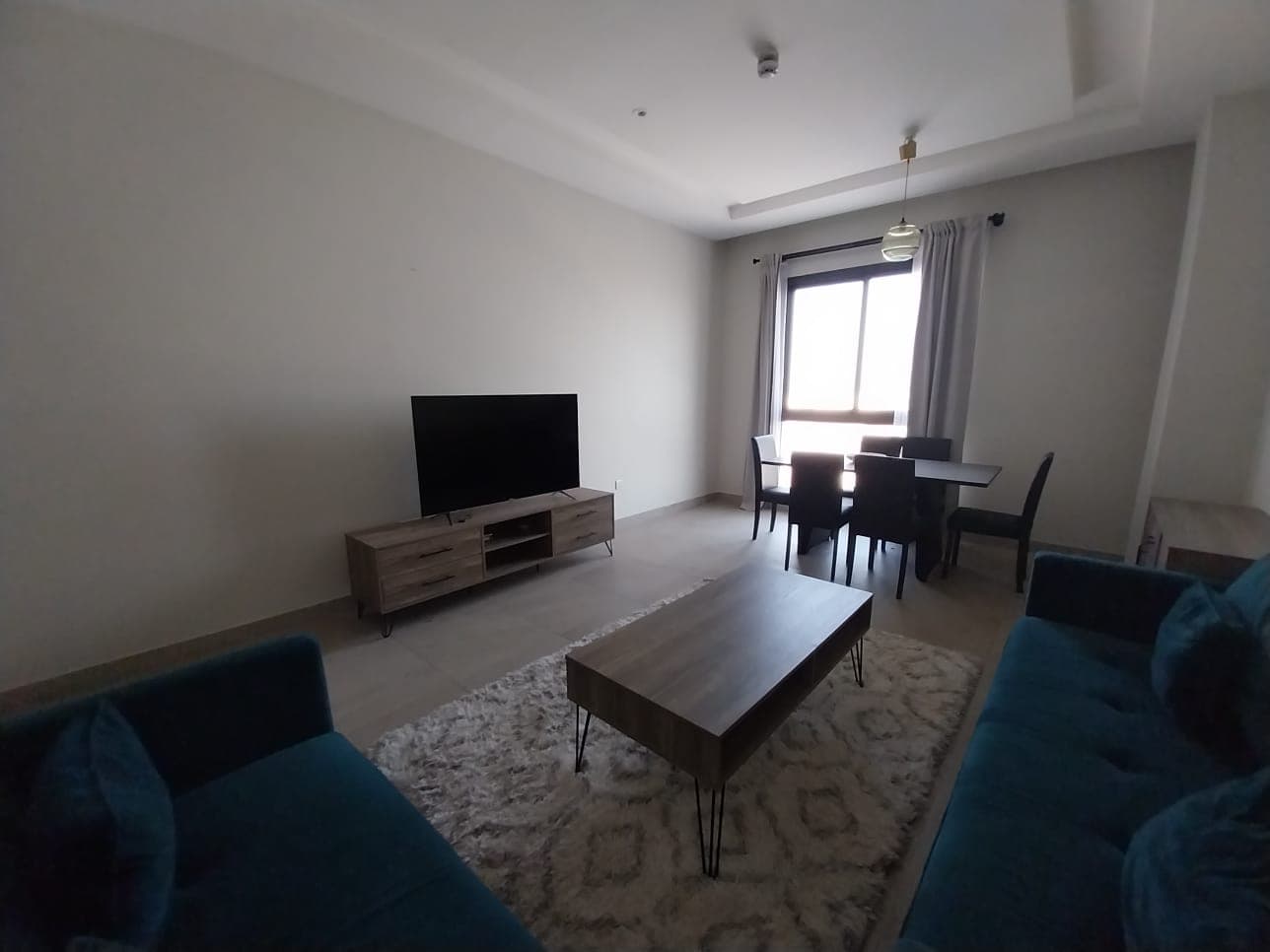 A flat for rent in Hidd features a cozy living room with two blue couches, a wooden coffee table on a rug, and a flat-screen TV on a stand. The space also includes a dining table with four chairs set in front of window curtains.