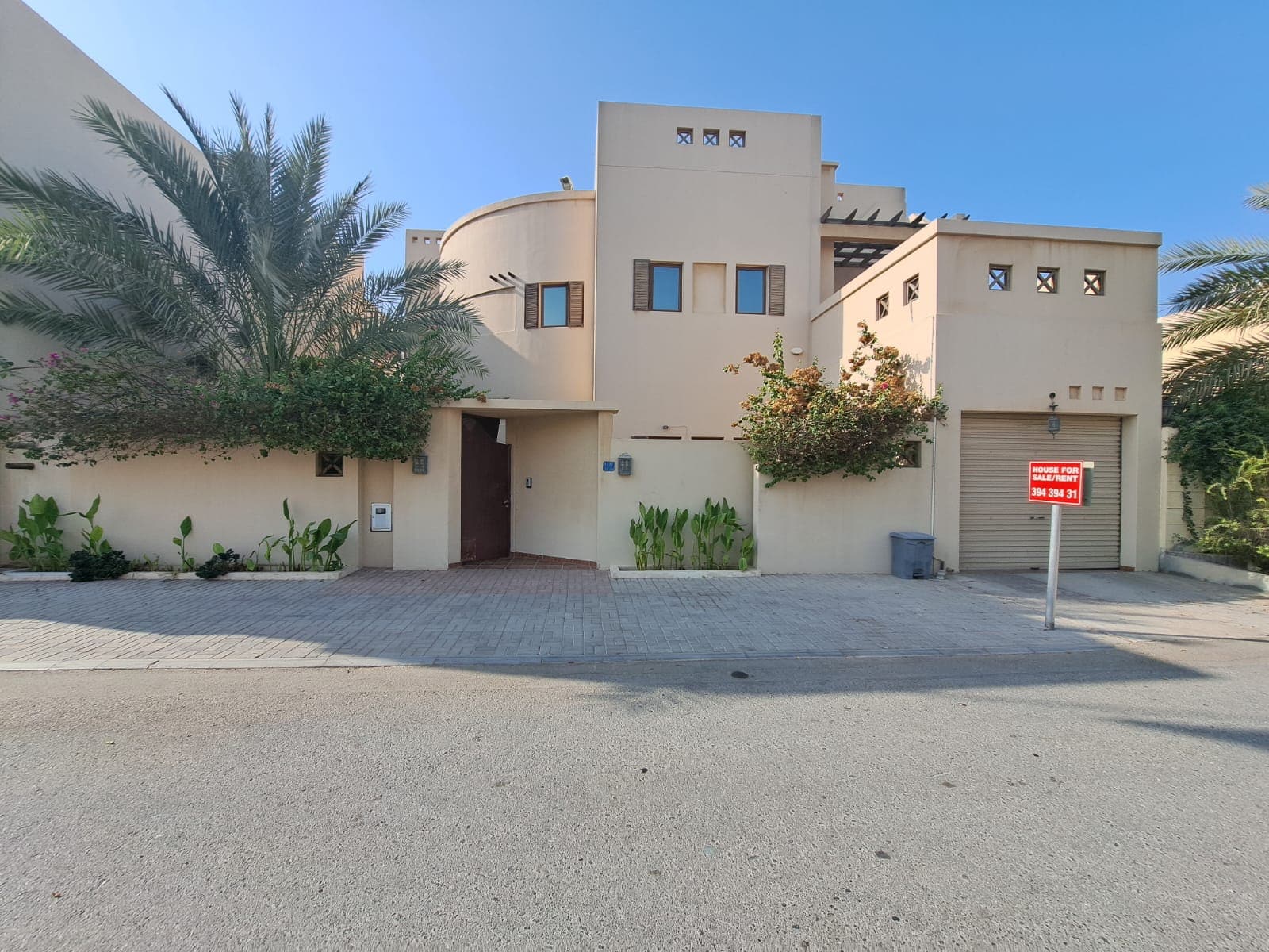 A modern beige villa with a curved design, surrounded by greenery. A "Villa for Sale" sign is posted near the driveway, inviting prospective homeowners to explore this elegant property.