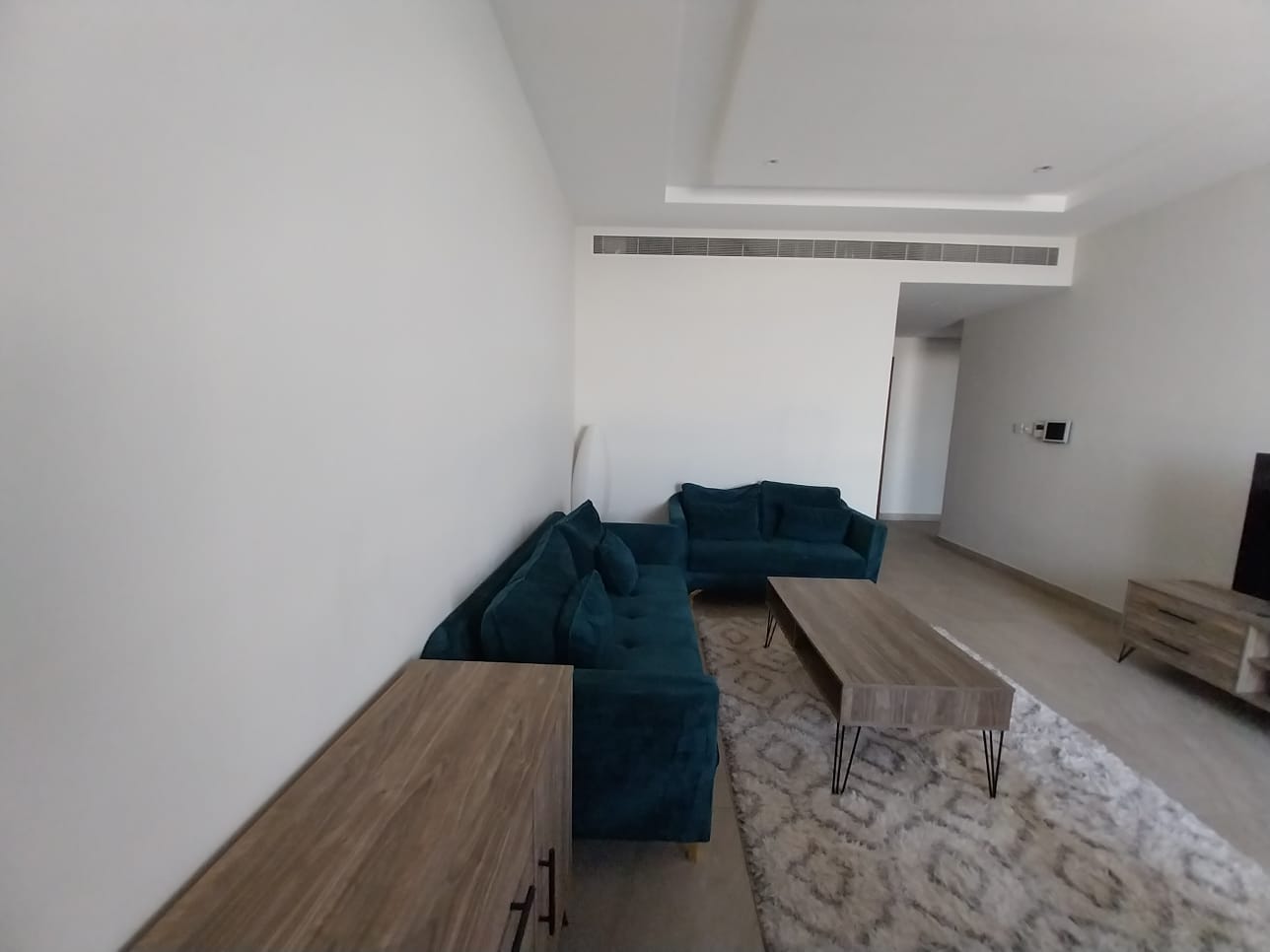 A minimalist living room with white walls, two teal sofas, a wooden coffee table, a small cabinet, and a patterned rug. An air vent is on the ceiling, and a console table is near the entrance. This flat for rent in Hidd offers modern comfort in every corner of its design.