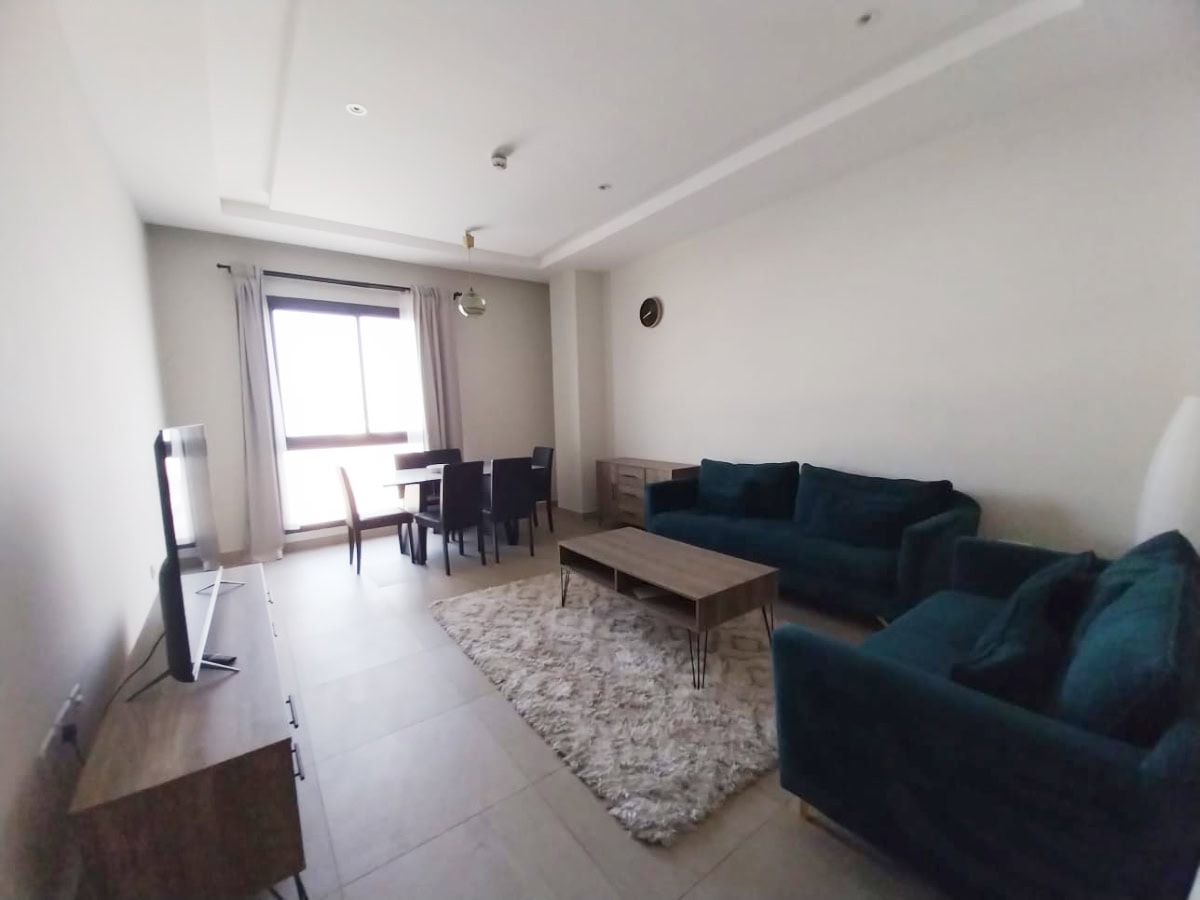 A modern living room in a Hidd flat for rent, featuring tile flooring, two dark green sofas, a wooden coffee table, a TV on a stand, a dining table with six chairs, and a large window with light curtains. Ideal as a cozy rental property.