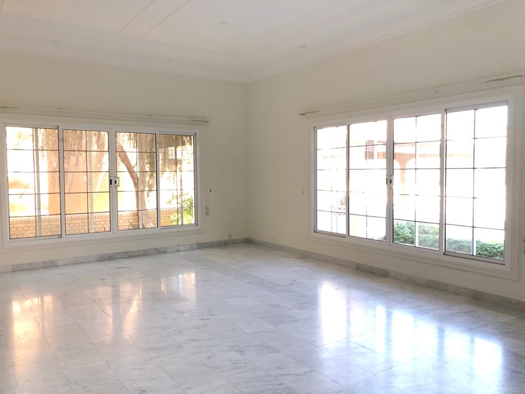 A semi-furnished, spacious room with large windows, white walls, and a light marble floor; outside greenery is visible through the windows.