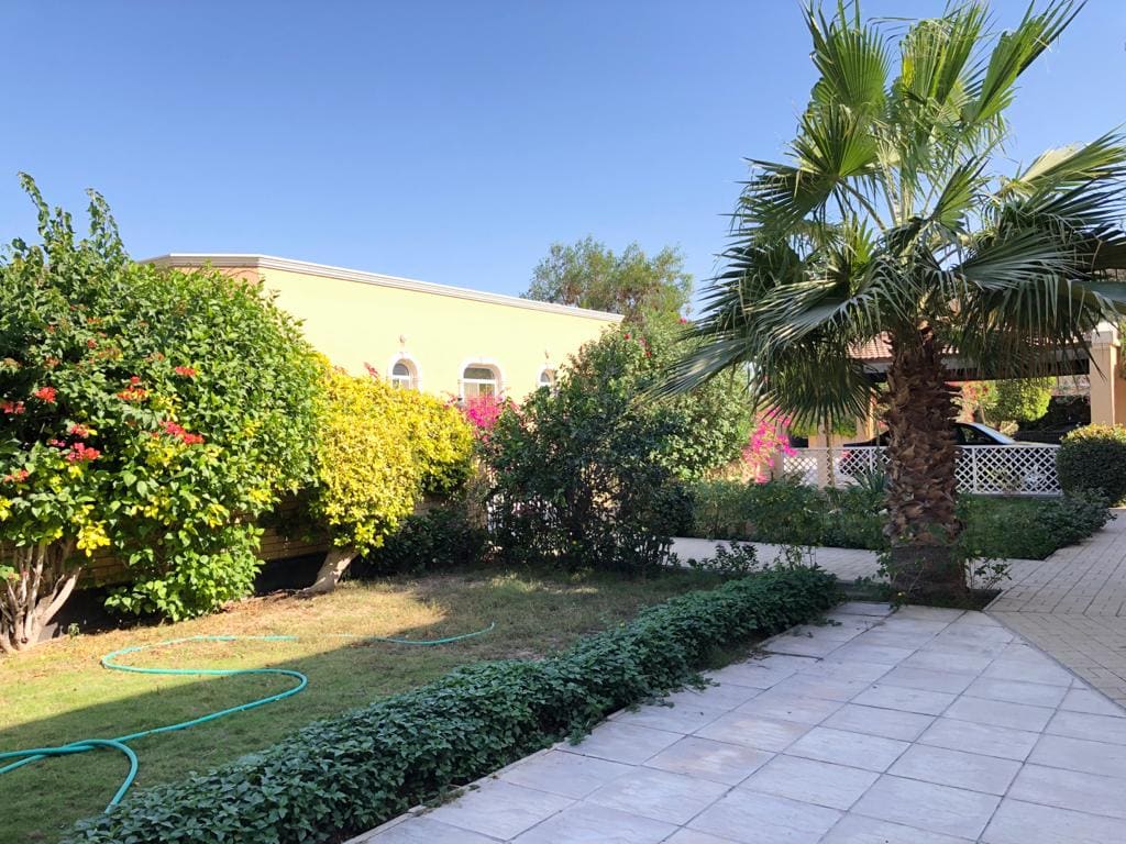 A spacious backyard garden with a tiled walkway, lush greenery, a palm tree, and shrubs. The yellow house, visible with arched windows and semi-furnished interiors, boasts 3 bedrooms. A coiled garden hose lies on the grass.