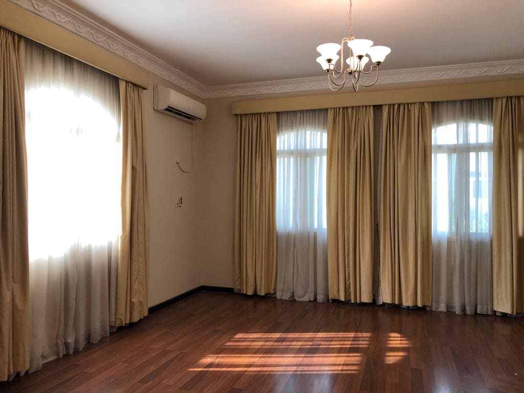 A 5-bedroom villa located in the Janbyah area, featuring a room with wooden flooring, three windows adorned with cream curtains, an air conditioner on the wall, and a ceiling light fixture. This semi-furnished property offers comfort and elegance.