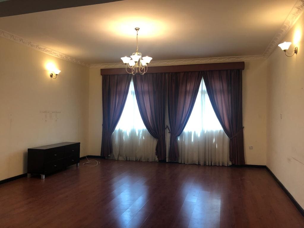 An elegant, semi-furnished room in a villa featuring hardwood flooring, two curtained windows, a chandelier, wall-mounted lights, and a dark cabinet against the wall.