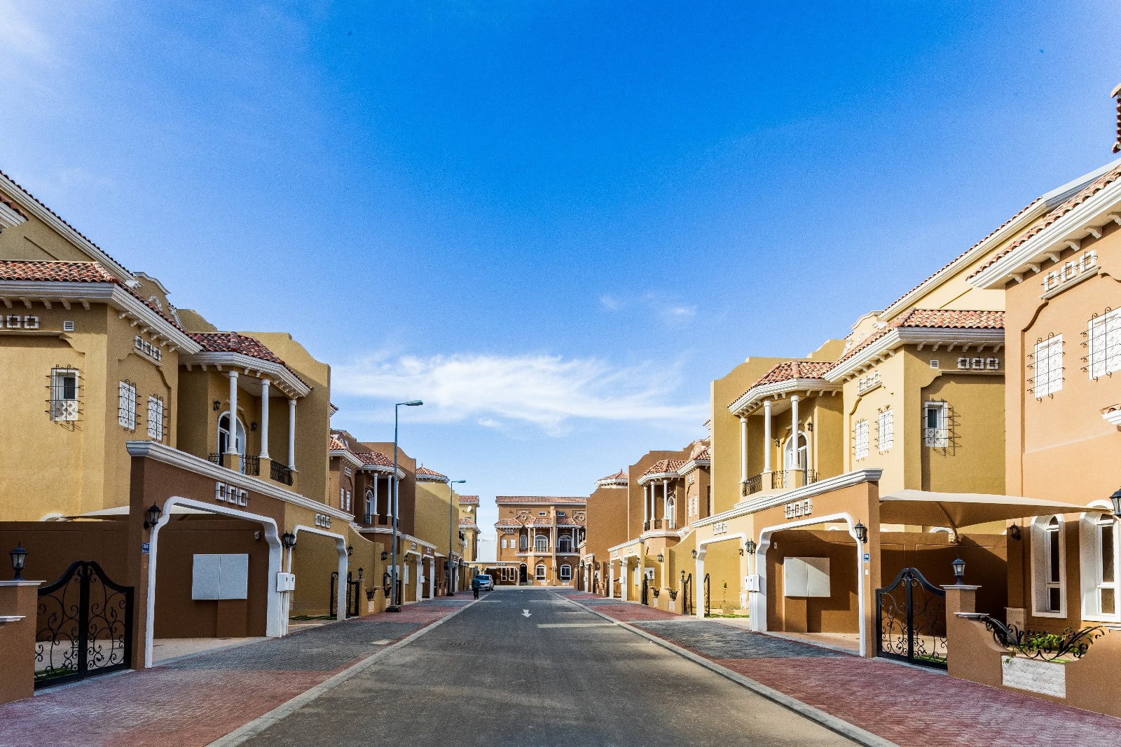 A luxury residential street in Janabiya features modern, multi-story villas in shades of yellow and orange under a clear blue sky. The road is flanked by well-maintained sidewalks and lampposts.