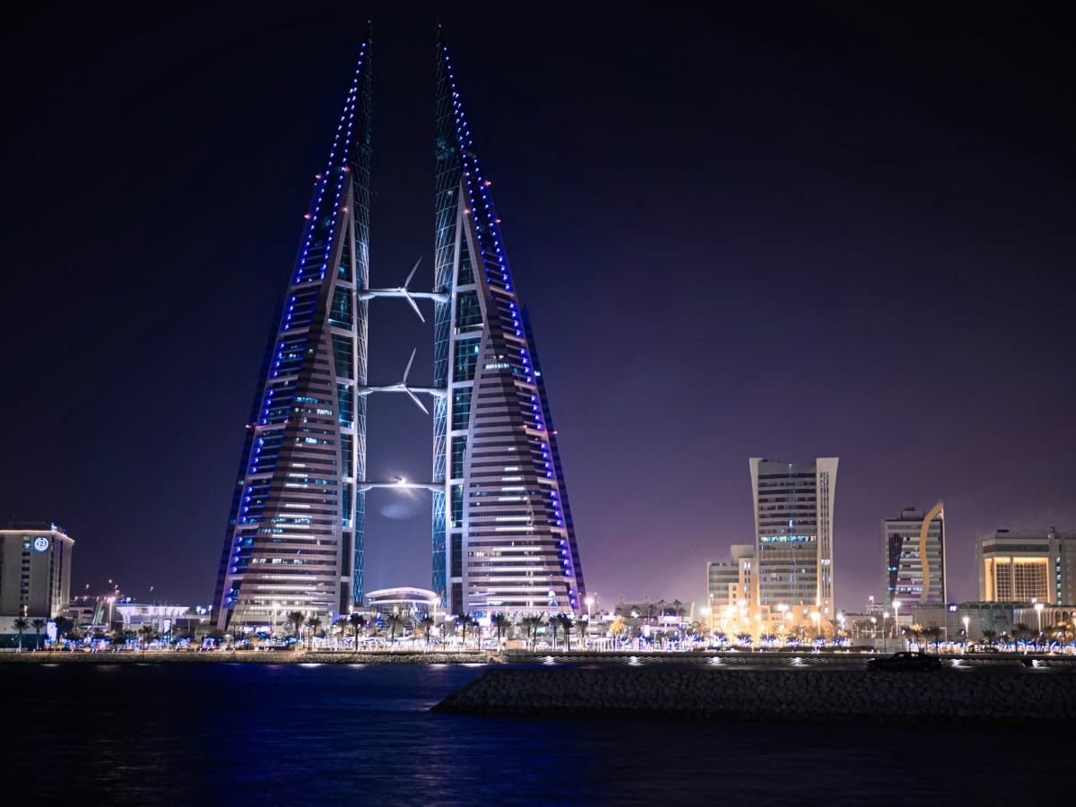 Night view of the illuminated Bahrain World Trade Center, a pair of sail-shaped towers with wind turbines, set against a backdrop of urban buildings and reflected on the water—a testament to Bahrain's thriving real estate market.