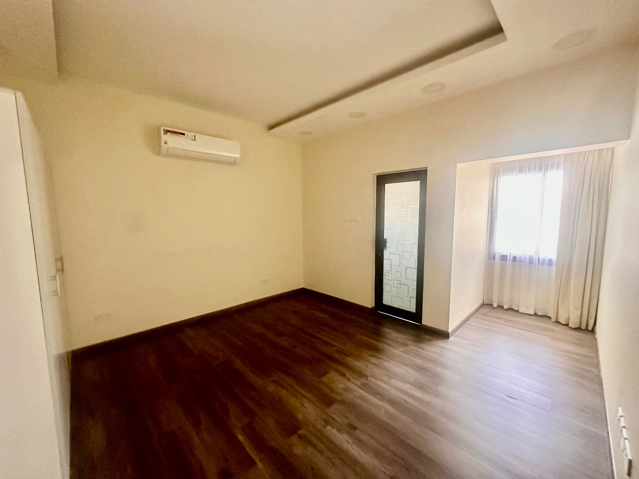 A vacant room with wooden flooring, white walls, an air conditioner, a large window with curtains, and a glass door with frosted design in a luxury villa in Janabiya.