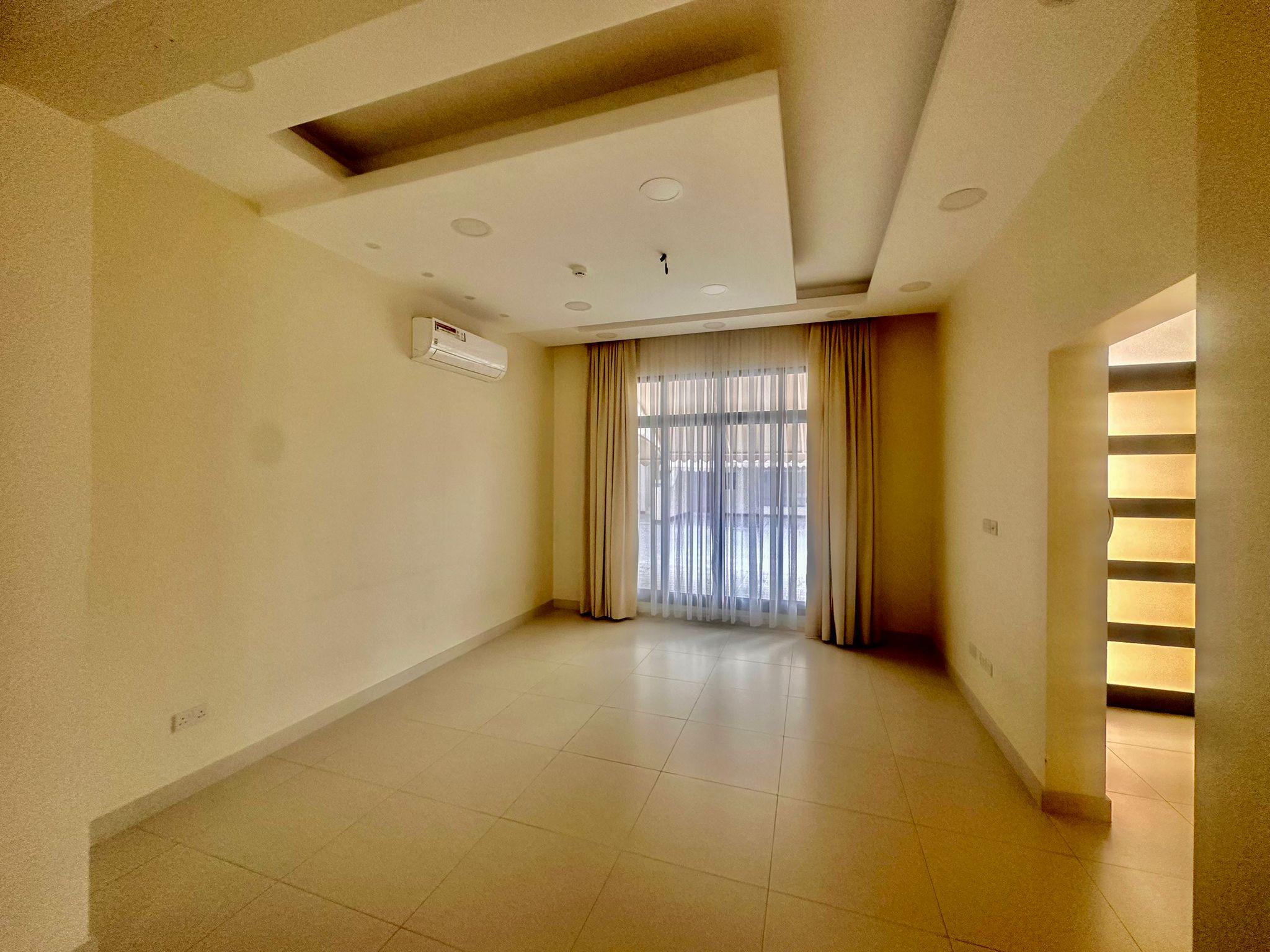 A spacious, empty room within a luxury villa in Janabiya, featuring tiled flooring, beige walls, ceiling lights, a window with sheer curtains, and an air conditioning unit mounted on the wall. This elegant room is part of a stunning 3-bedroom residence.