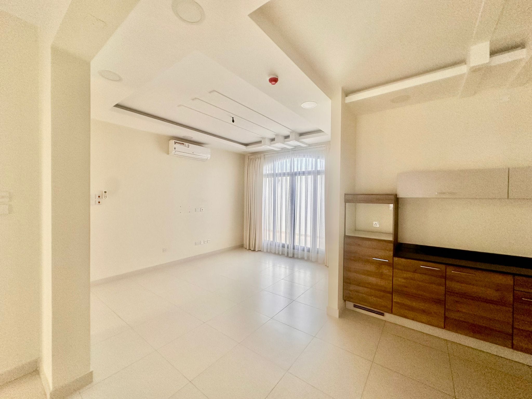 A spacious, empty room with white tiled floors, ceiling lights, air conditioner, large window with sheer curtains, and a wooden kitchenette on the right is part of this luxury villa in Janabiya featuring 3 bedrooms.