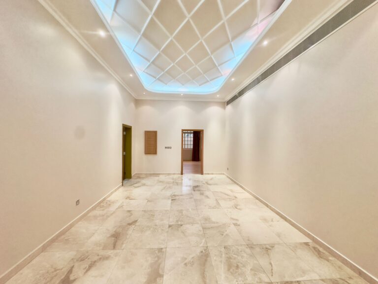 Spacious room with white walls, marble tile flooring, and a patterned ceiling featuring recessed lighting and a skylight. Located in a 2 bedrooms villa within the Al Janabiyah compound, two open doorways are visible on the left side.