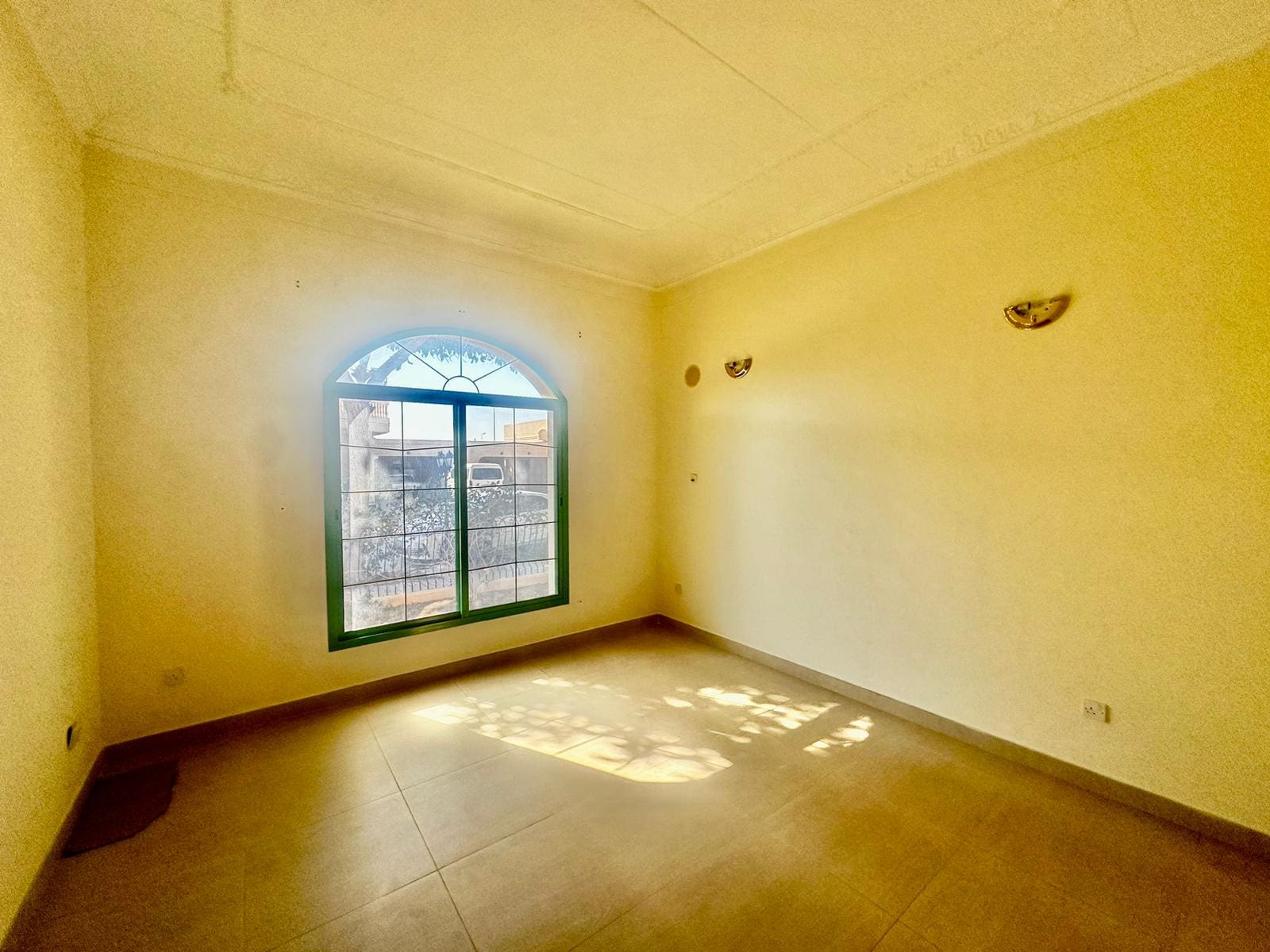 An empty room with beige walls, a large arched window, tile flooring, and two wall-mounted light fixtures sits in the semi-furnished 3 bedrooms villa. Sunlight streaming through the window casts shadows on the floor in this serene Janbyah area home.