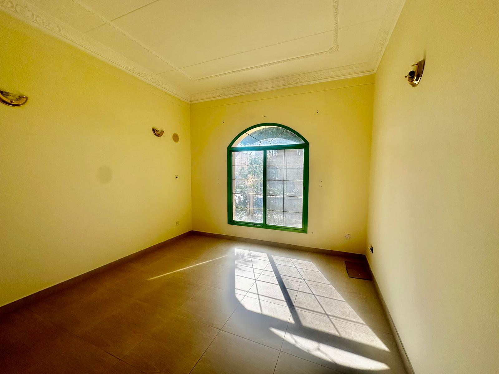 An empty room with cream-colored walls, a tiled floor, and an arched window allowing sunlight to enter. Wall-mounted lights are on both sides of the window in this semi-furnished villa located in the Janbyah area.