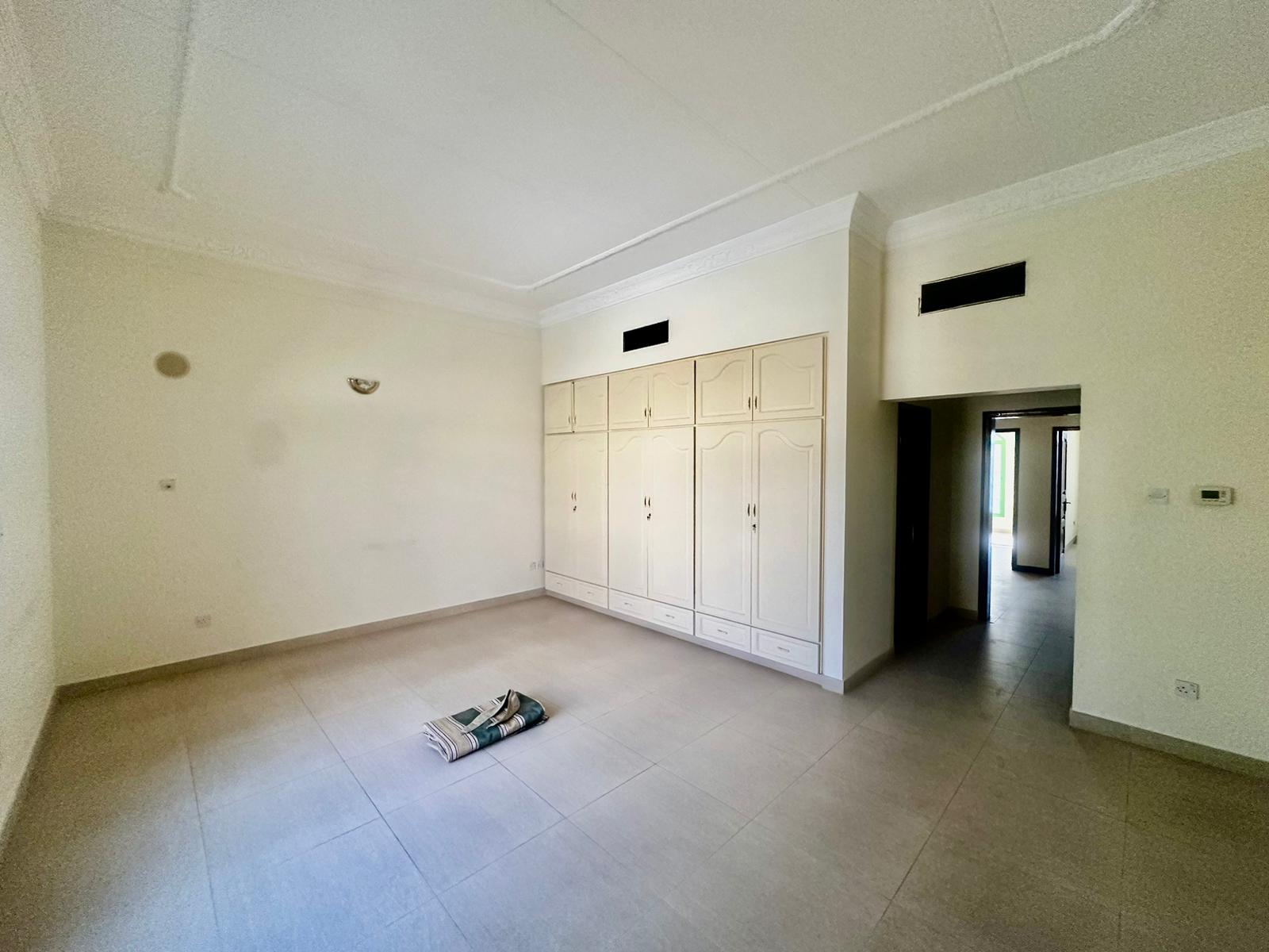 A spacious, semi-furnished room with light-colored walls and floor tiles in a charming 3 bedrooms villa in the Janbyah area. There is a built-in wardrobe on one side and a rolled rug in the center. An open doorway leads to another room.