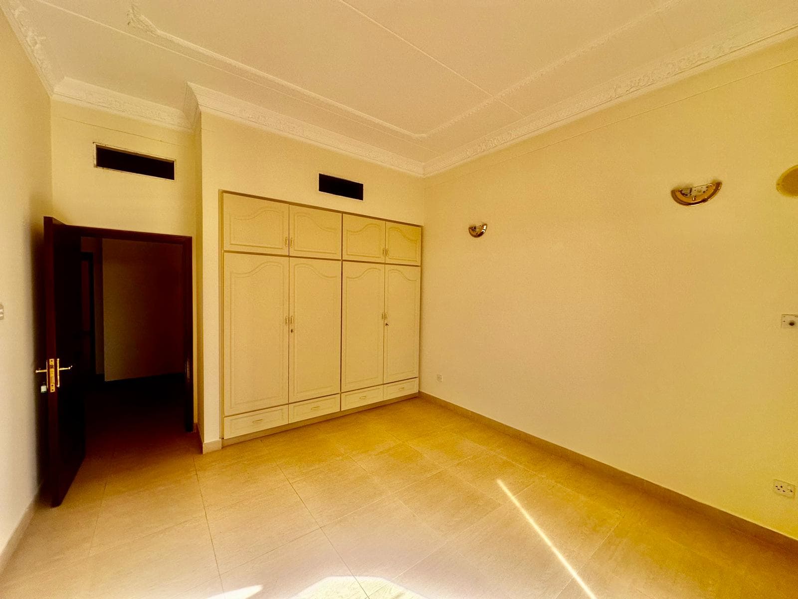 A semi-furnished room with a beige wardrobe, light-colored walls, tiled floor, two small wall-mounted lights, and a wooden door slightly ajar lies within the spacious 3 bedrooms villa.