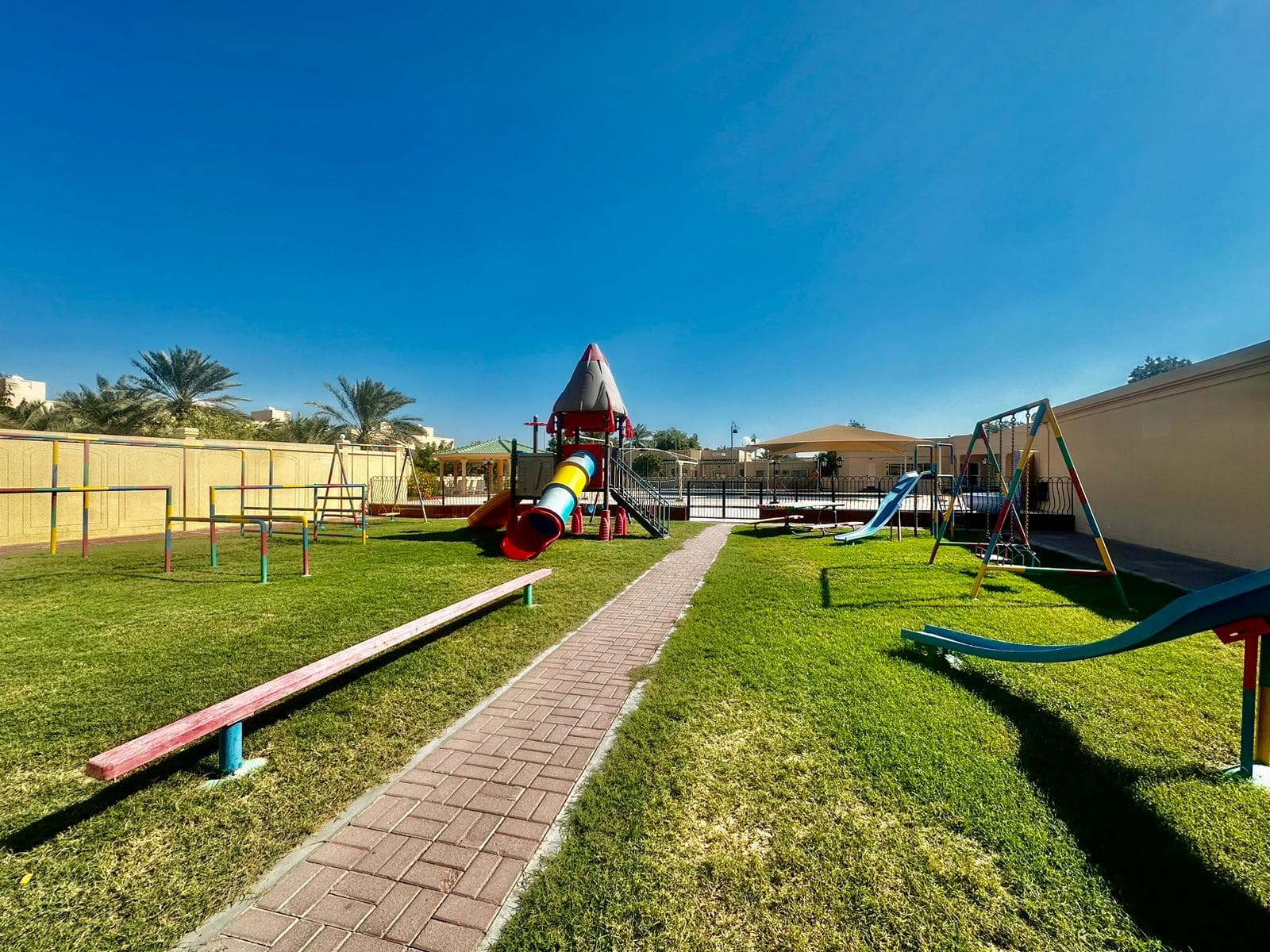 A playground with a slide, swings, a rocket-shaped climbing structure, and balancing beams on a grassy area under a clear blue sky is adjacent to a charming 3-bedroom villa in the Janbyah area.