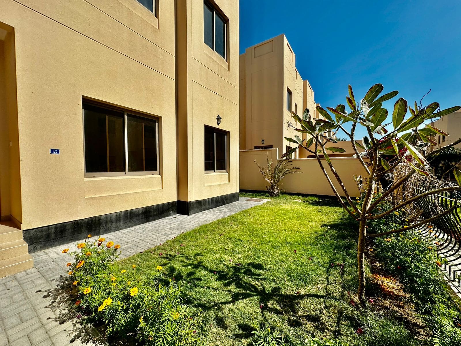 A beige villa with large windows overlooks a small, neatly maintained garden with green grass, flowering plants, and a bare tree. Located in Janbiyah, this beautiful property offers 4 bedrooms. Above, the sky is clear and blue.