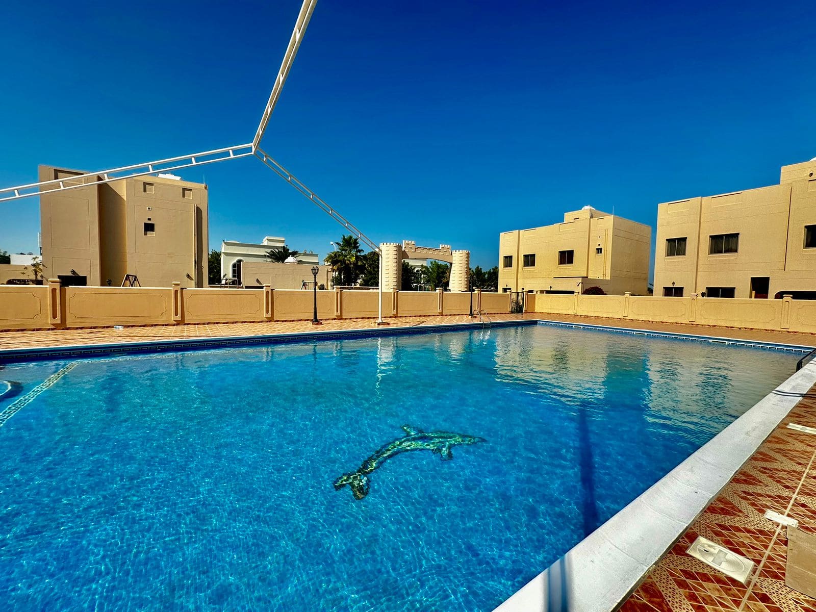 Outdoor swimming pool surrounded by beige buildings under a clear blue sky, with a dolphin design at the bottom of the pool in this stunning 4-bedroom villa in Janbiyah.