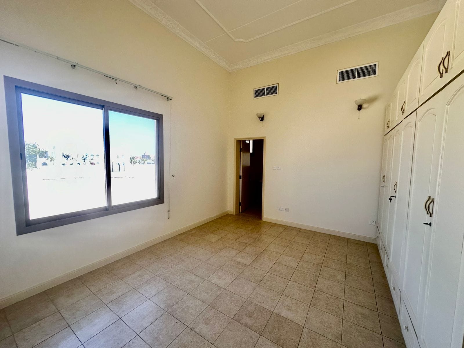 A vacant room with a tiled floor, a large window on the left wall, white built-in wardrobes on the right, and a door in the middle of the back wall sits within this 4 bedrooms villa compound in Janbiyah area. Ceiling lights and vents are also visible.