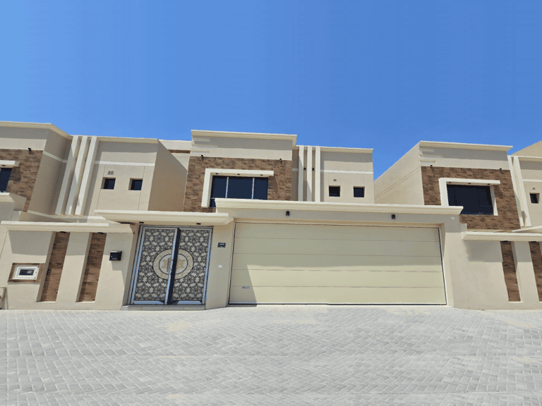 A contemporary beige two-story house for sale in the Damistan area, featuring a double garage, modern design elements, and a distinctive decorative entry door under a clear blue sky. One of the stunning new villas in this sought-after neighborhood.