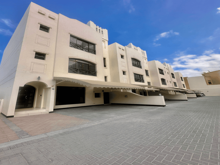 A row of modern beige apartment buildings with arched entrances and overhangs, situated on a paved area with parked vehicles, evokes the elegance of a luxury villa in Janabiya. The sky is partly cloudy, adding a serene backdrop to the 3-bedroom residences.