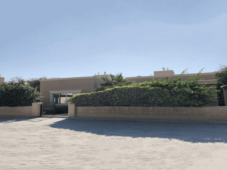 A spacious, single-story 3-bedroom villa with a flat roof, surrounded by a brick wall and dense green bushes, under a clear blue sky.