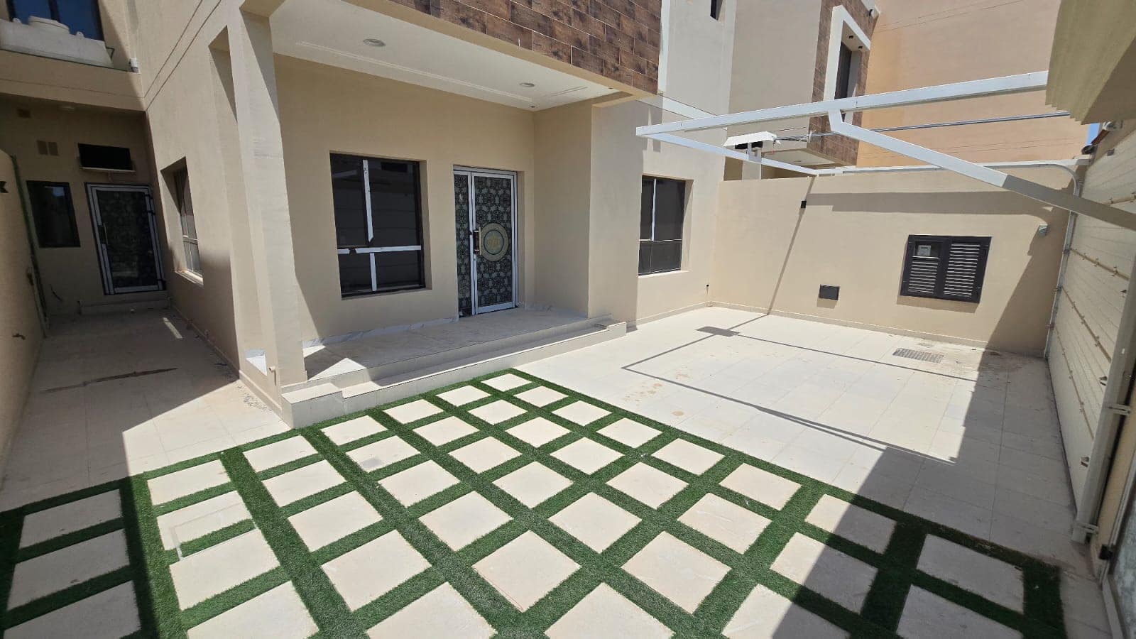 A modern house with a beige exterior features a tiled patio with grass strips, windows, and a decorative front door. Located in Damistan, this new villa offers a yard with a mix of pavement and grassy patches and is currently for sale.