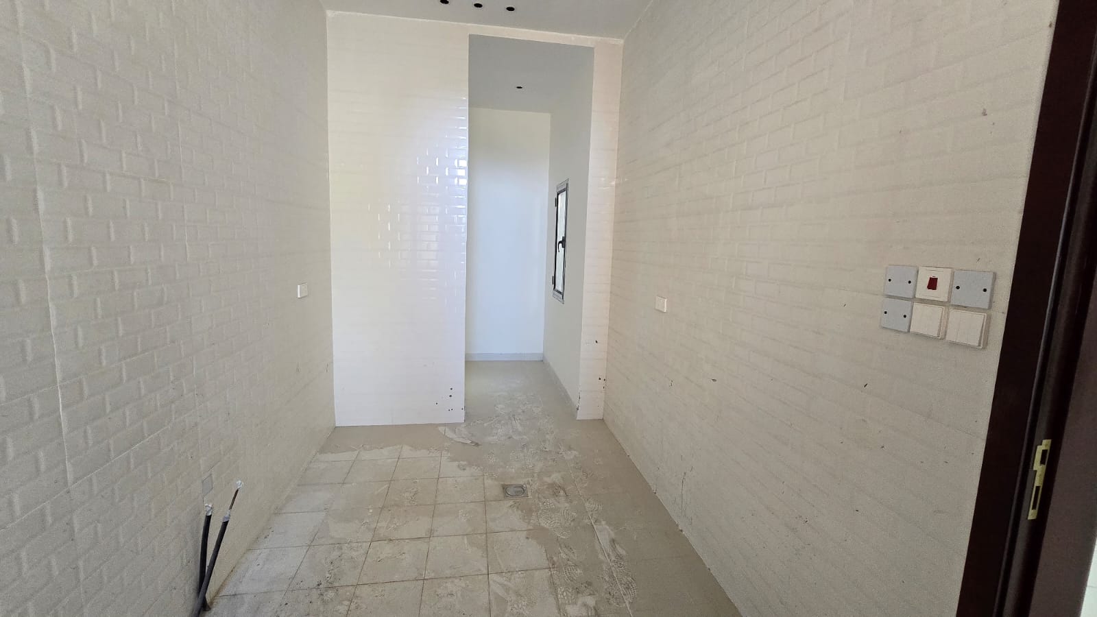 A narrow, empty room with white tiled walls and a tiled floor. Several electrical outlets are on the right wall. There is a narrow doorway at the end of the room. Positioned in one of Damistan's new villas for sale, this space offers flexibility and modern comfort.