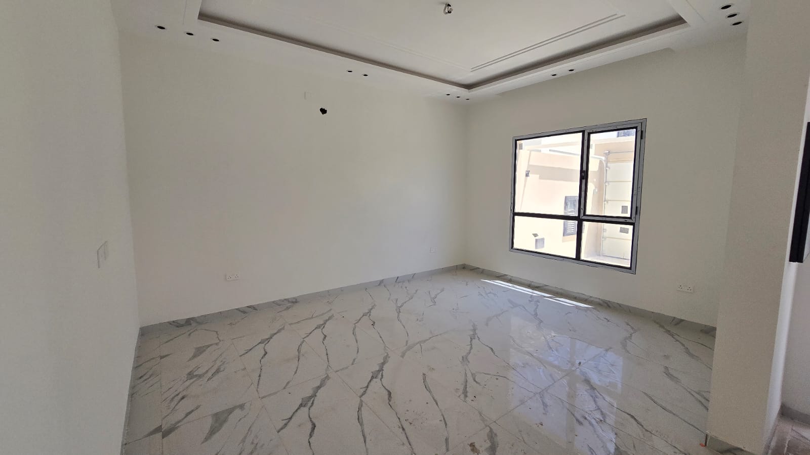 Empty room with white walls, a large window, and marble tile flooring. The ceiling features recessed lighting spaces without fixtures installed. Sunlight enters through the window. Ideal for new villas in Damistan, this space is now for sale.