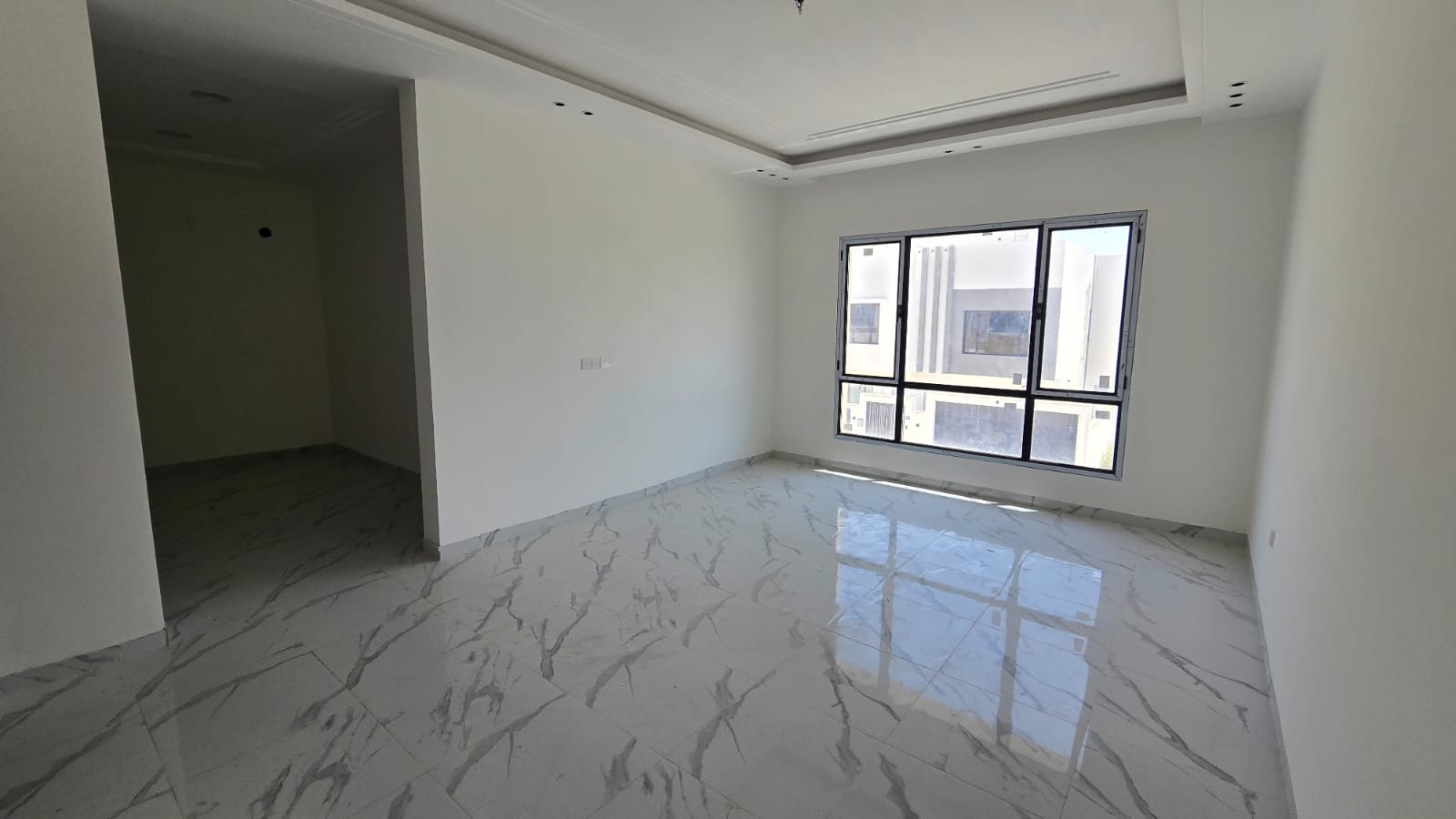 A vacant, unfurnished room with marble floor tiles, white walls, a large window, and a doorway leading to another area in one of the new villas for sale in Damistan.