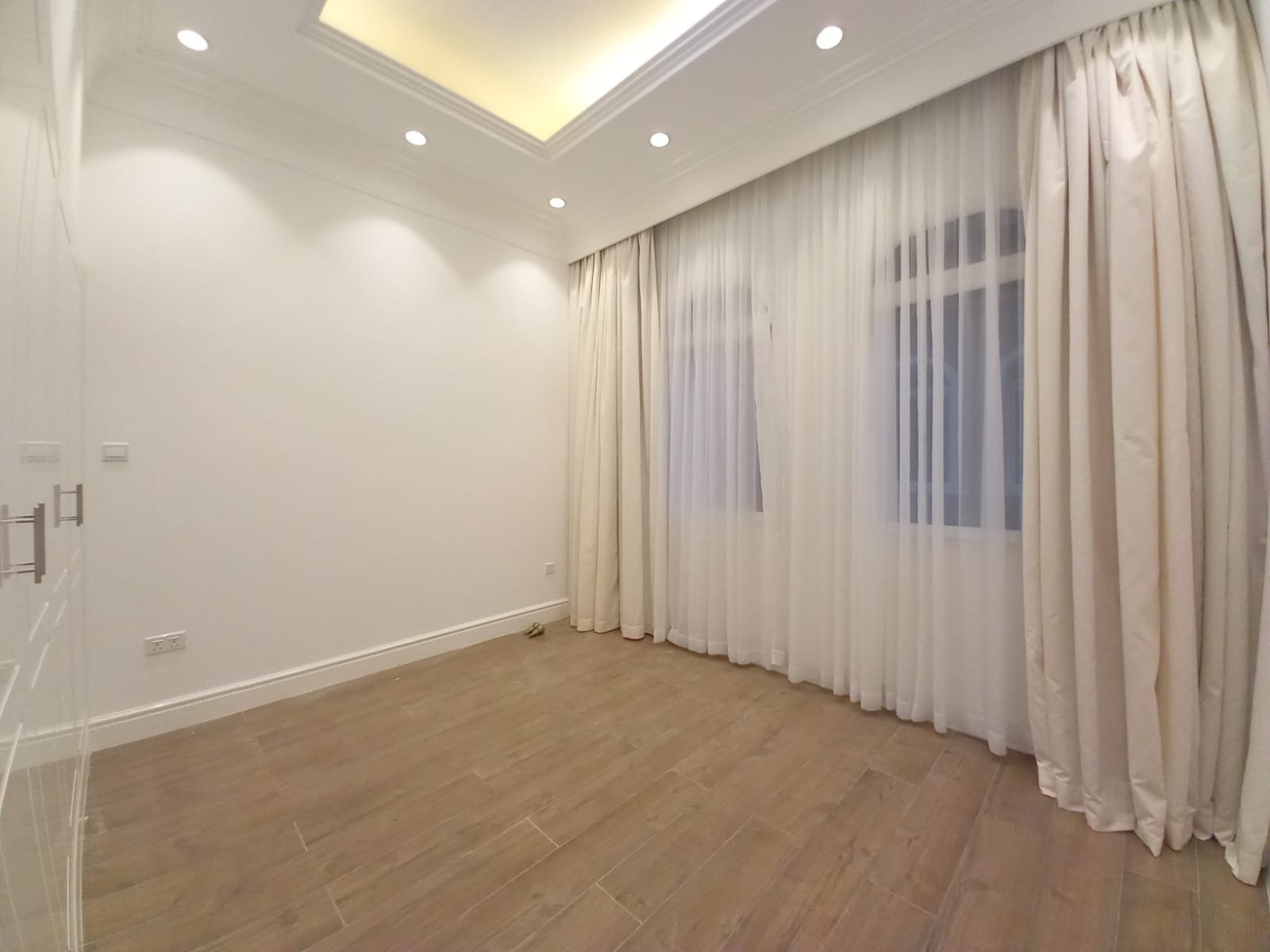 A well-lit empty room with white walls, recessed ceiling lights, wooden floor, and large windows covered by beige curtains in a luxury villa in Janabiya with 3 bedrooms.