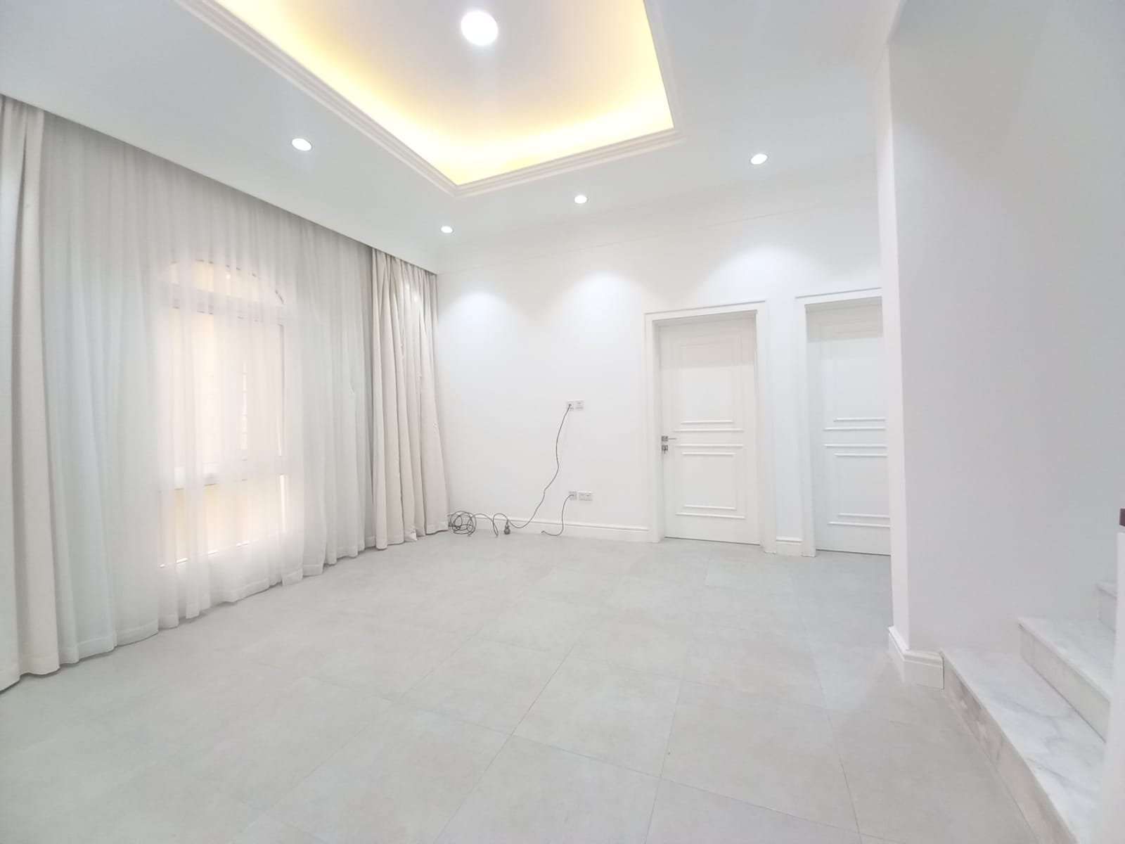 A minimalist room in a luxury villa for rent in Janabiya, featuring white walls, a recessed ceiling with lights, floor-to-ceiling curtains, two closed white doors, and some electrical wires on the floor.