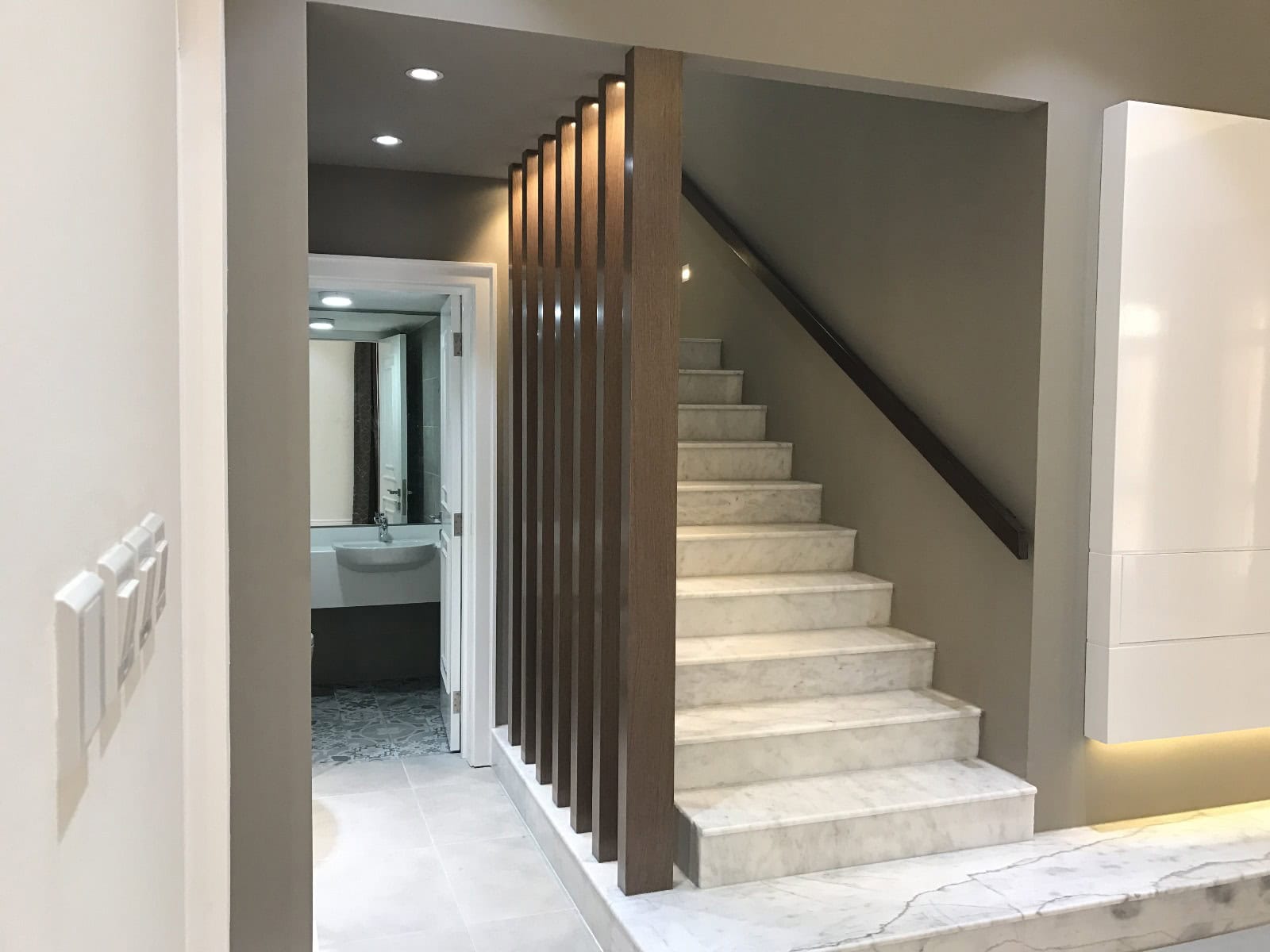 Modern interior featuring a white marble staircase with wooden slats, leading up to the next floor in this luxury villa. A bathroom can be seen at the end of a hallway to the left of the stairs in this stunning Janabiya home that boasts 3 bedrooms.