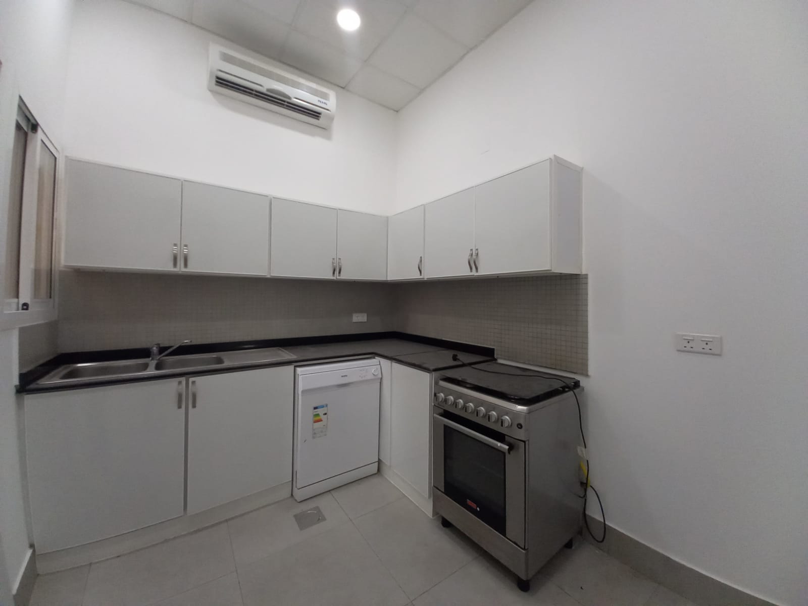 A small, modern kitchen in a luxury villa with white cabinets, black countertops, a stainless steel sink, a dishwasher, a gas stove, and white walls. An air conditioning unit is mounted above the cabinets. This elegant space complements the villa's 3 bedrooms located in Janabiya.